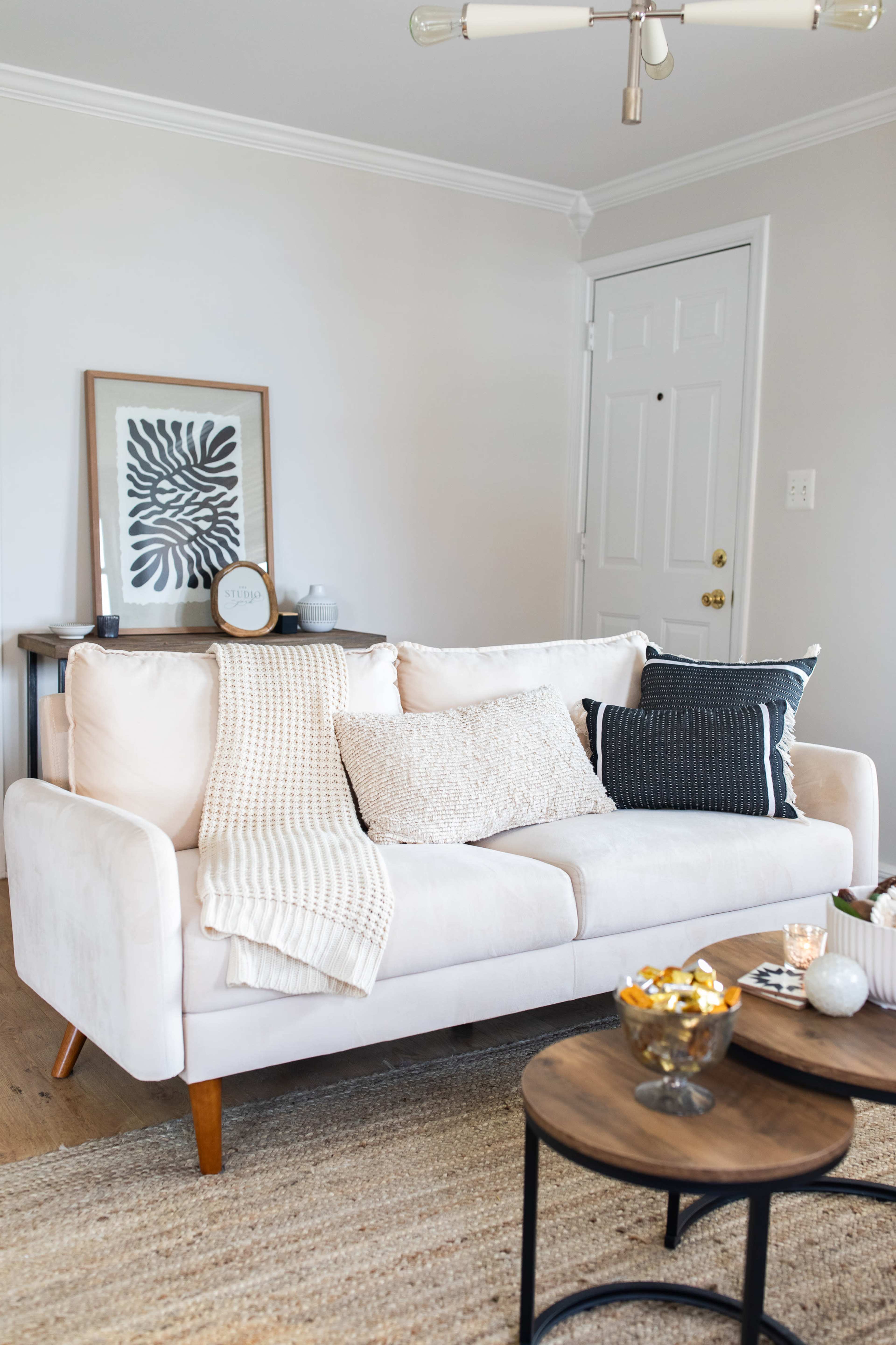 A light-colored sofa with various decorative pillows and a knitted throw blanket is positioned in a living room with wooden floors and a small coffee table.