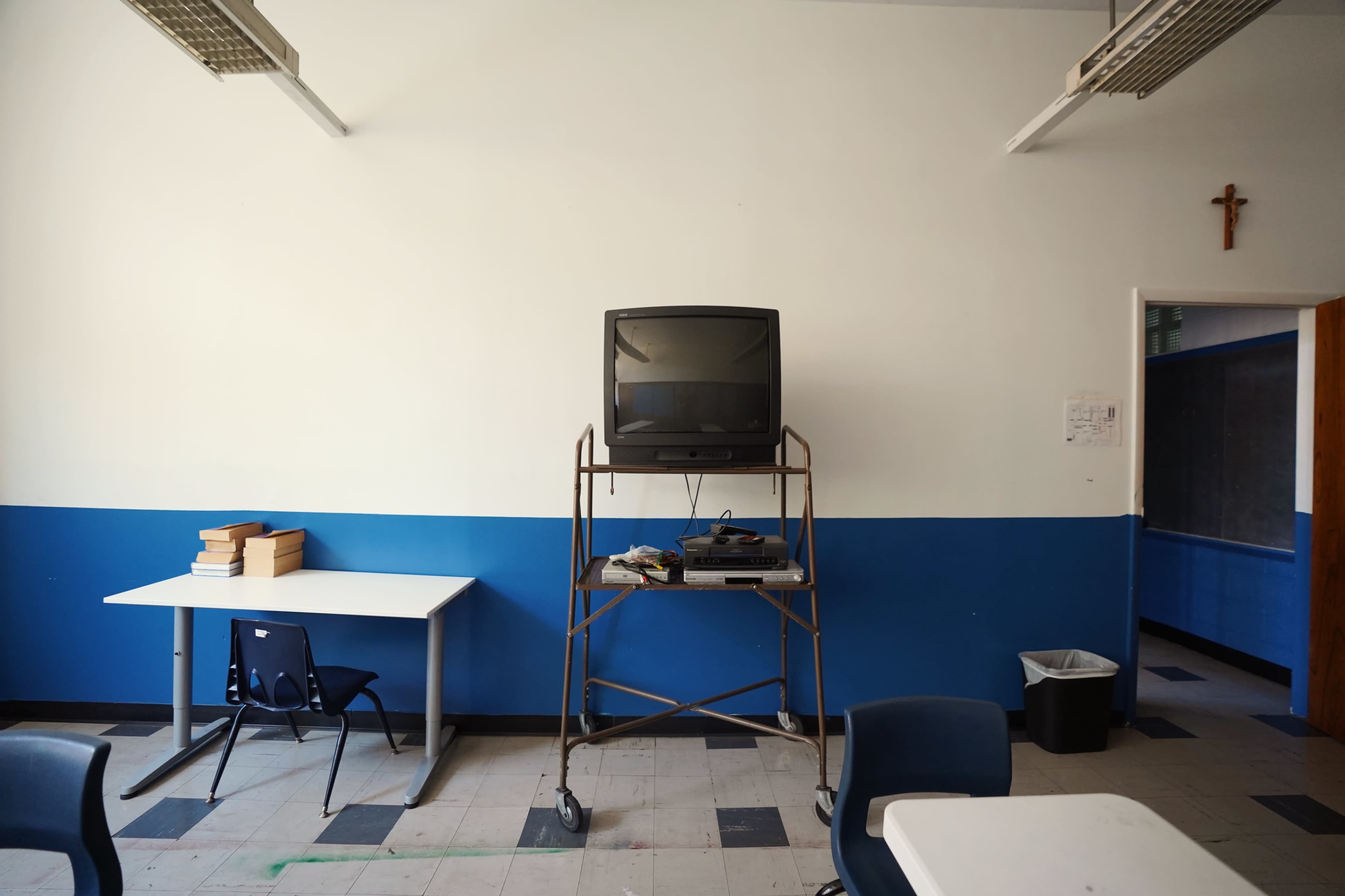 A classroom with a television on a metal stand, positioned against a blue and white wall, accompanied by a stack of books on a table and a crucifix on the wall.