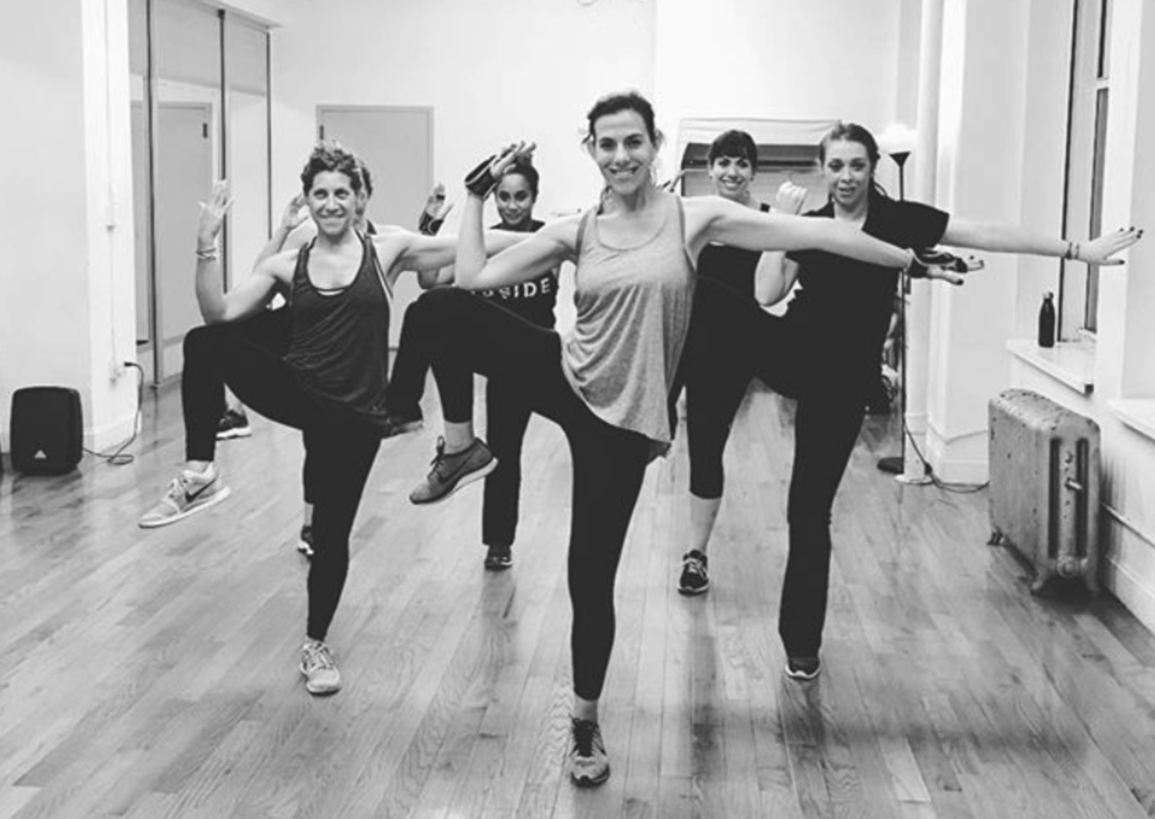A group of six women is performing a dance routine in a studio with wooden floors and large windows.
