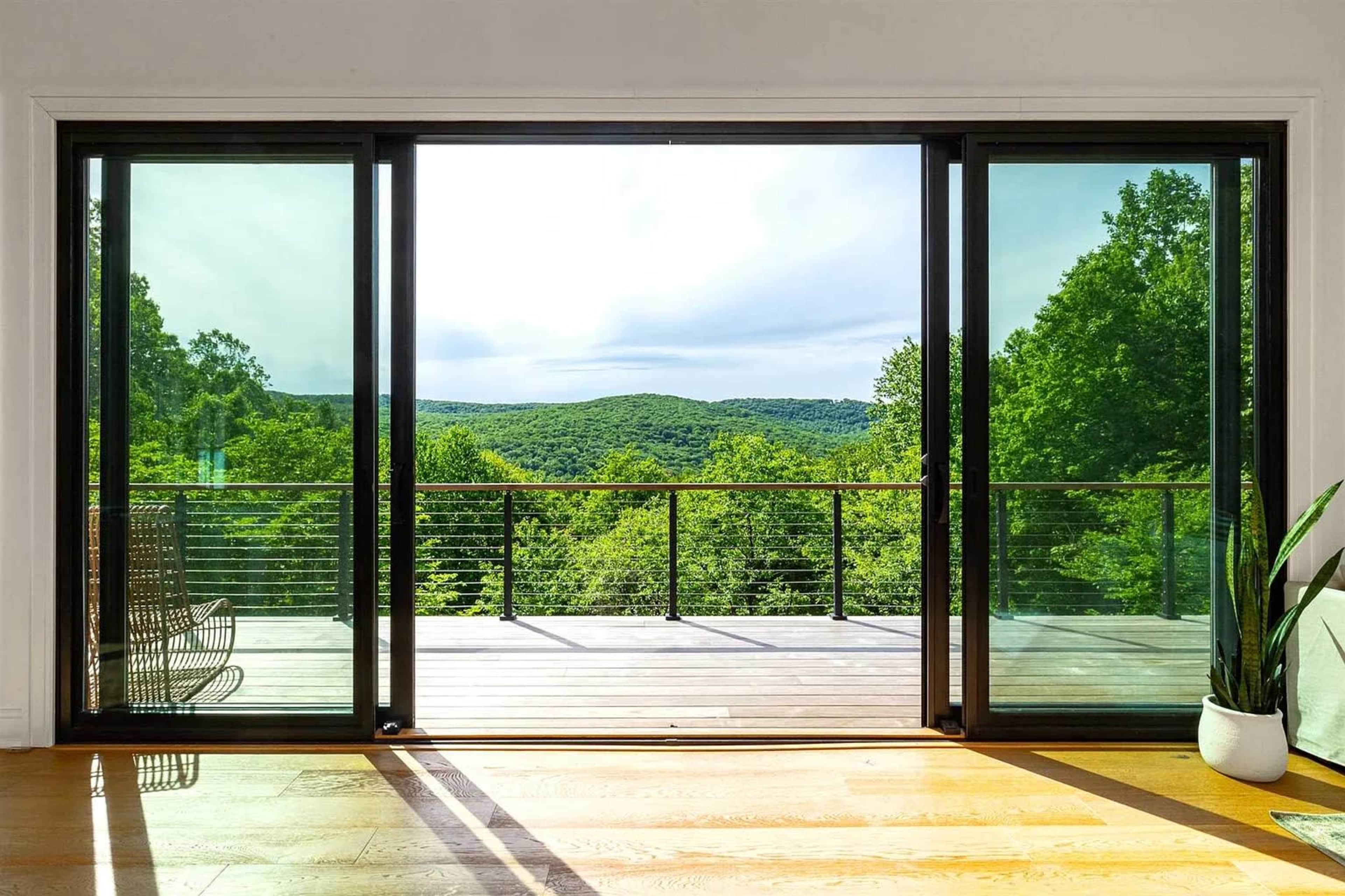 A spacious deck with glass sliding doors offers a view of lush green hills under a cloudy sky.