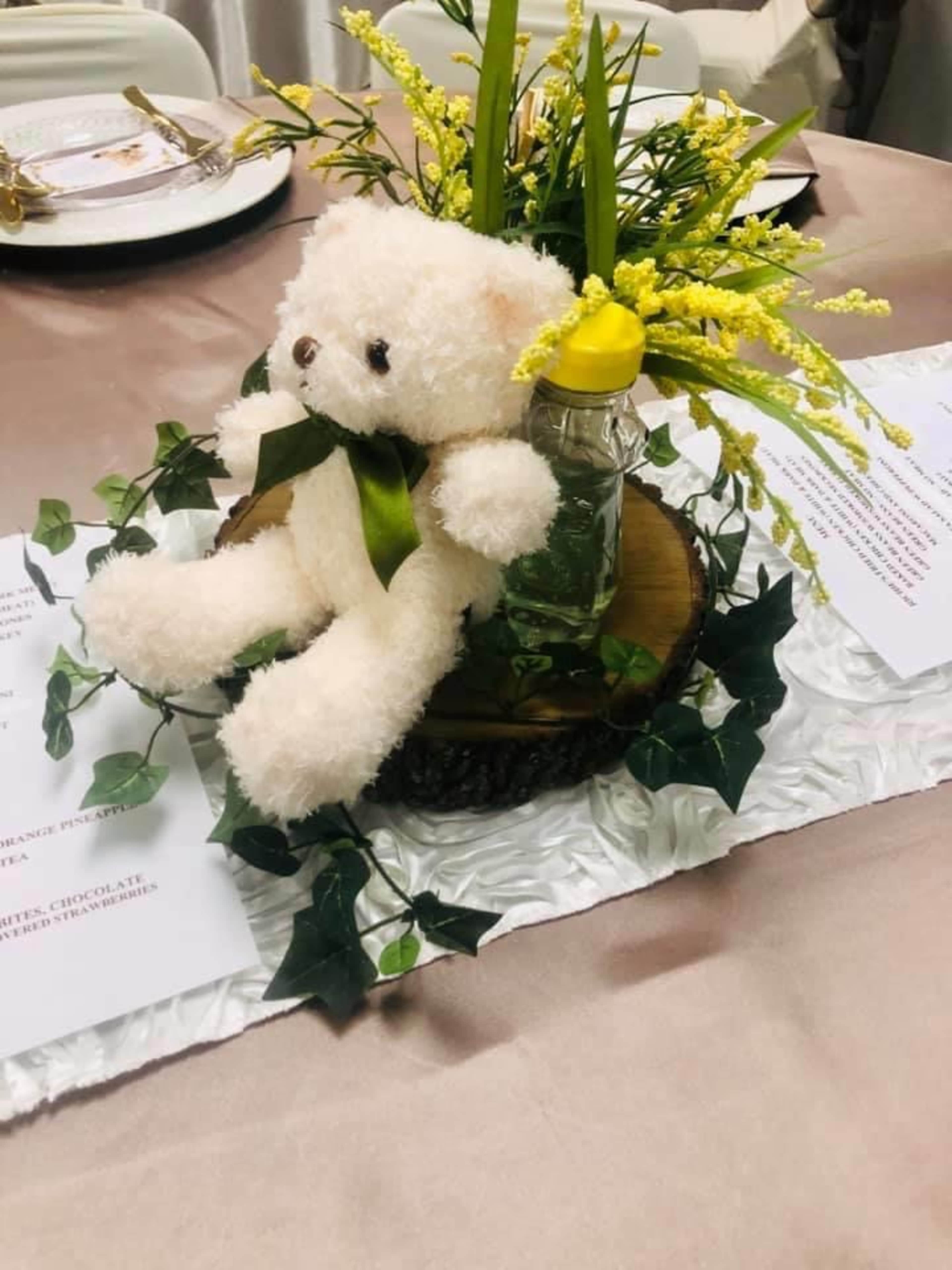 A plush teddy bear sits on a wooden slice with a glass jar filled with flowers, surrounded by green ivy on a decorated table.