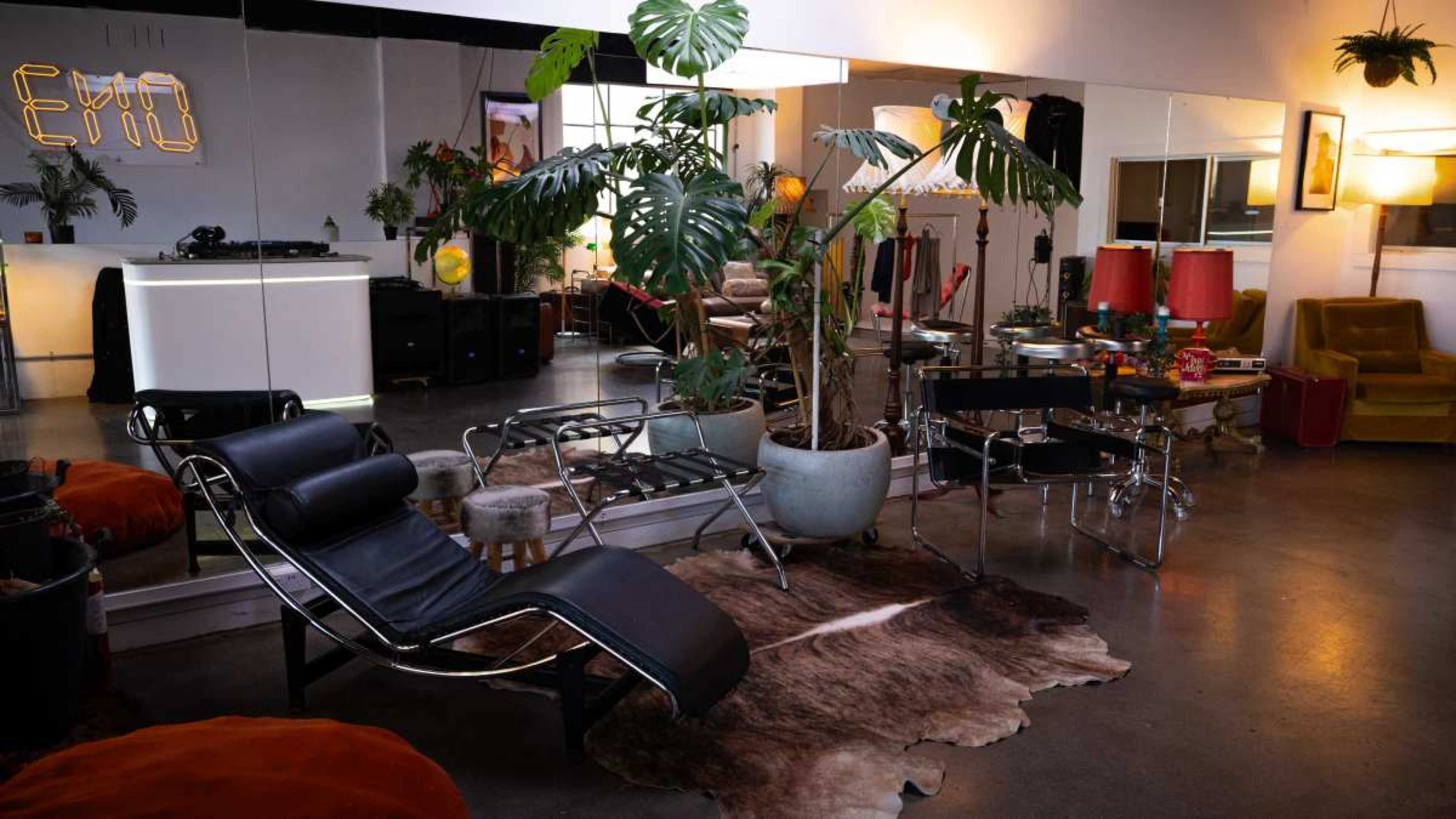 The image depicts a stylish interior space featuring modern furniture, including a black lounge chair, a glass-top table, and various indoor plants, with a mirrored wall reflecting the decor.