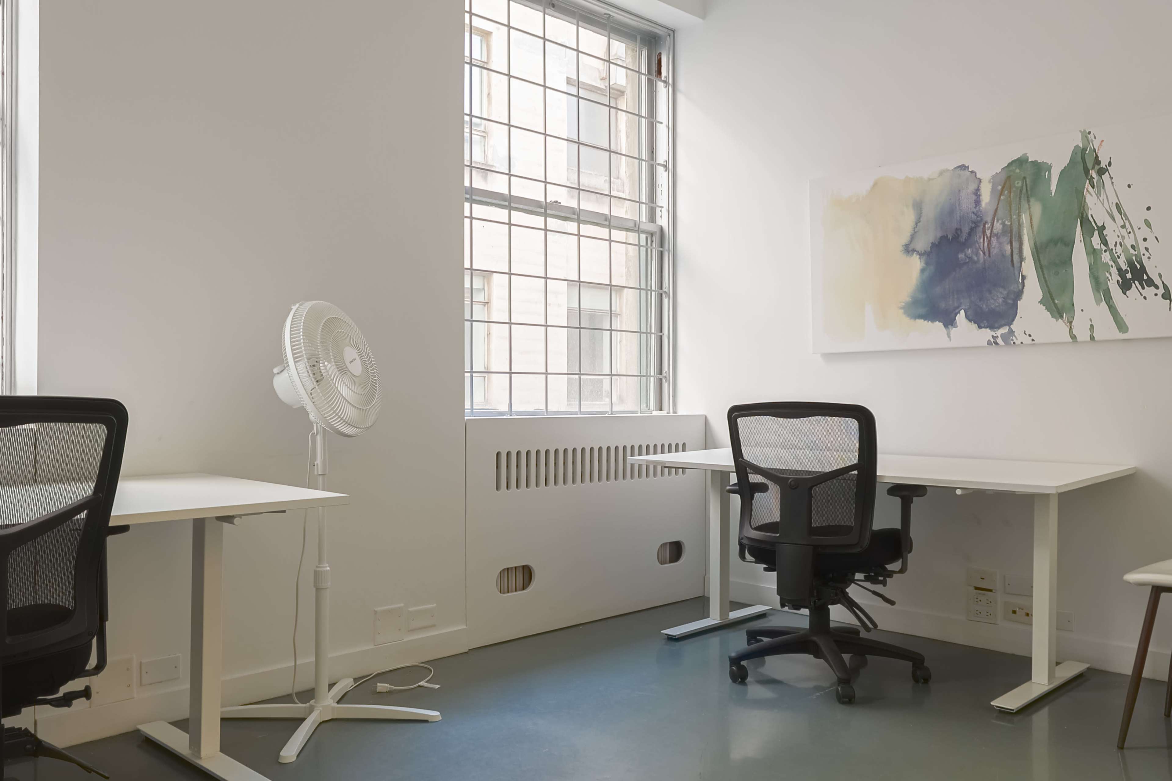 The image shows an empty office space featuring two desks, a fan, and an abstract wall painting with large windows.