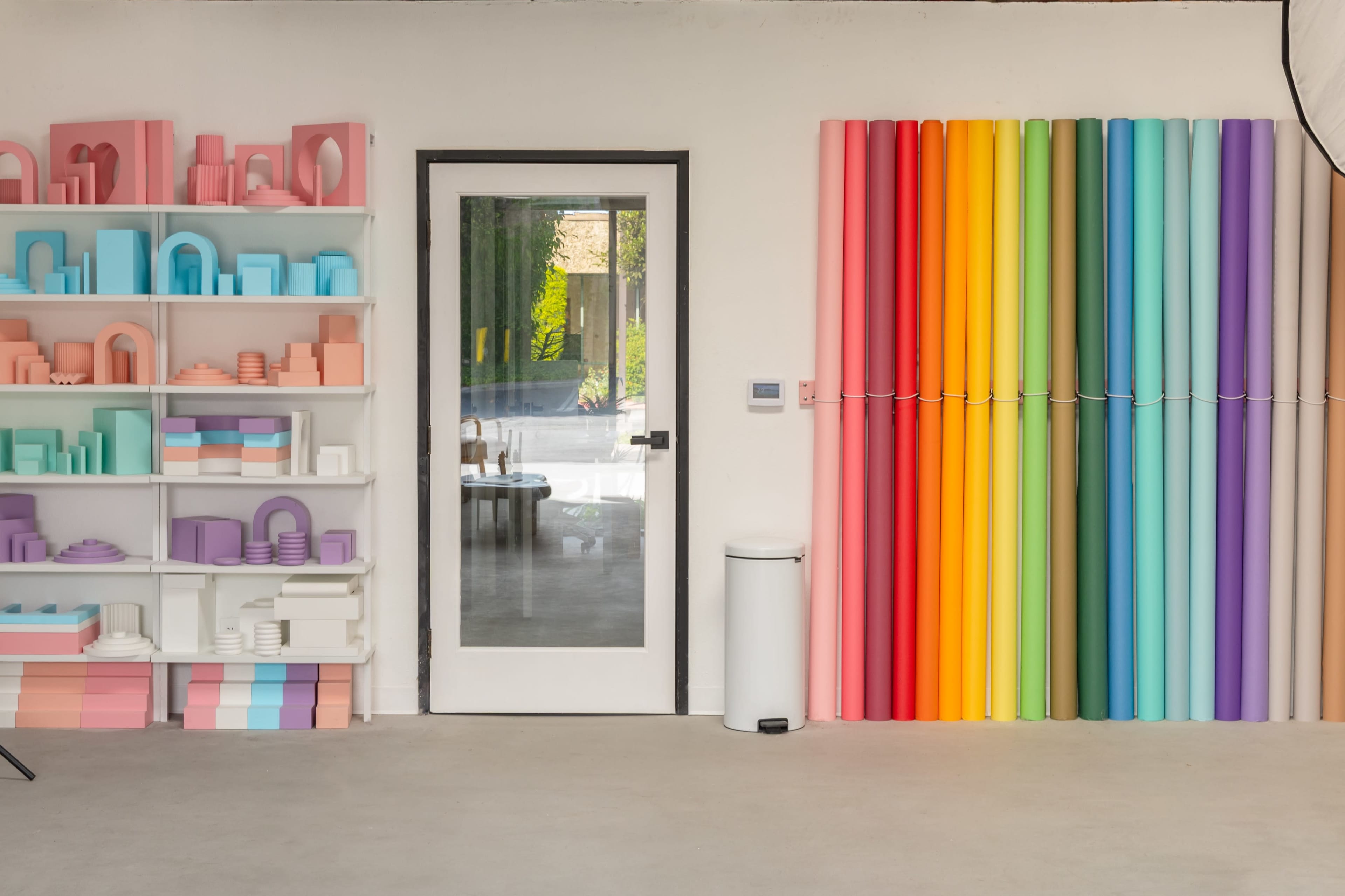 The image shows a modern interior with a white wall featuring colorful rolled paper and a shelf displaying various geometric shapes in pastel colors, adjacent to a glass door.