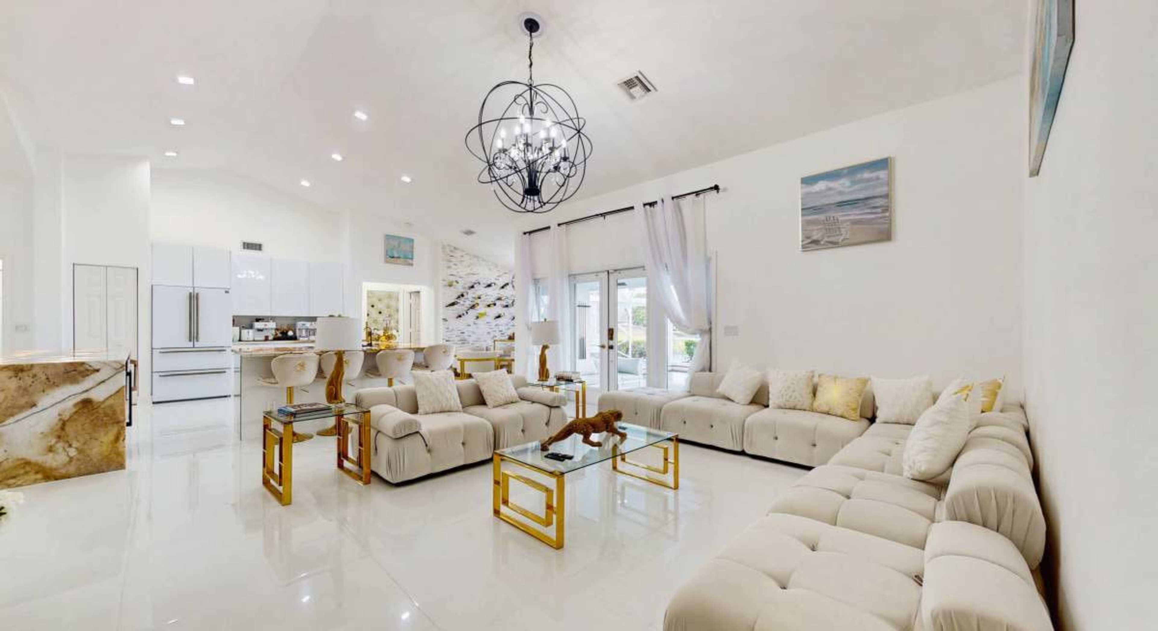 The image shows a modern living room featuring a large sectional sofa, a glass coffee table, and a decorative chandelier, with large windows allowing natural light to enter.