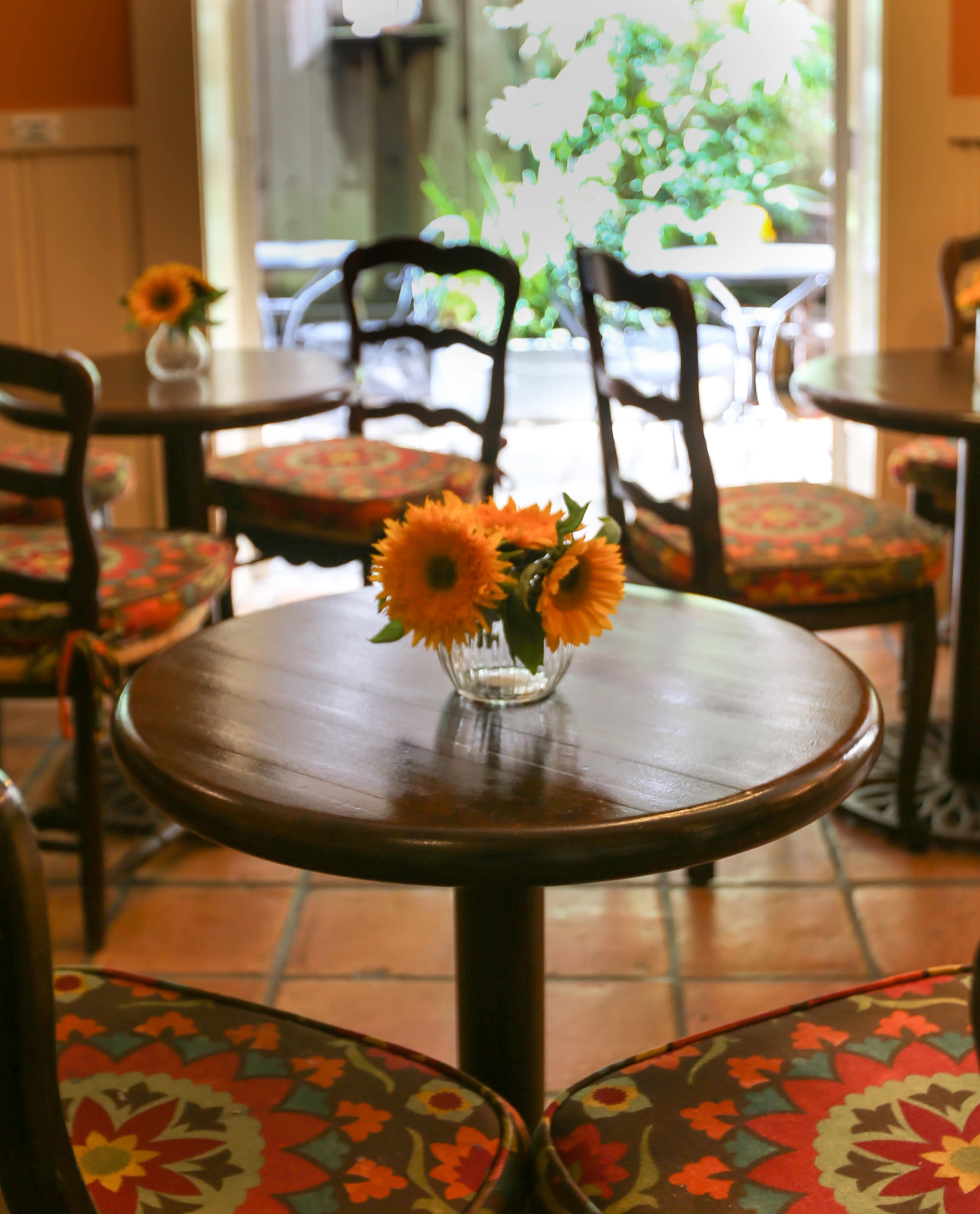 A small wooden table with a vase of sunflowers is surrounded by chairs in a cafe with an outdoor view.