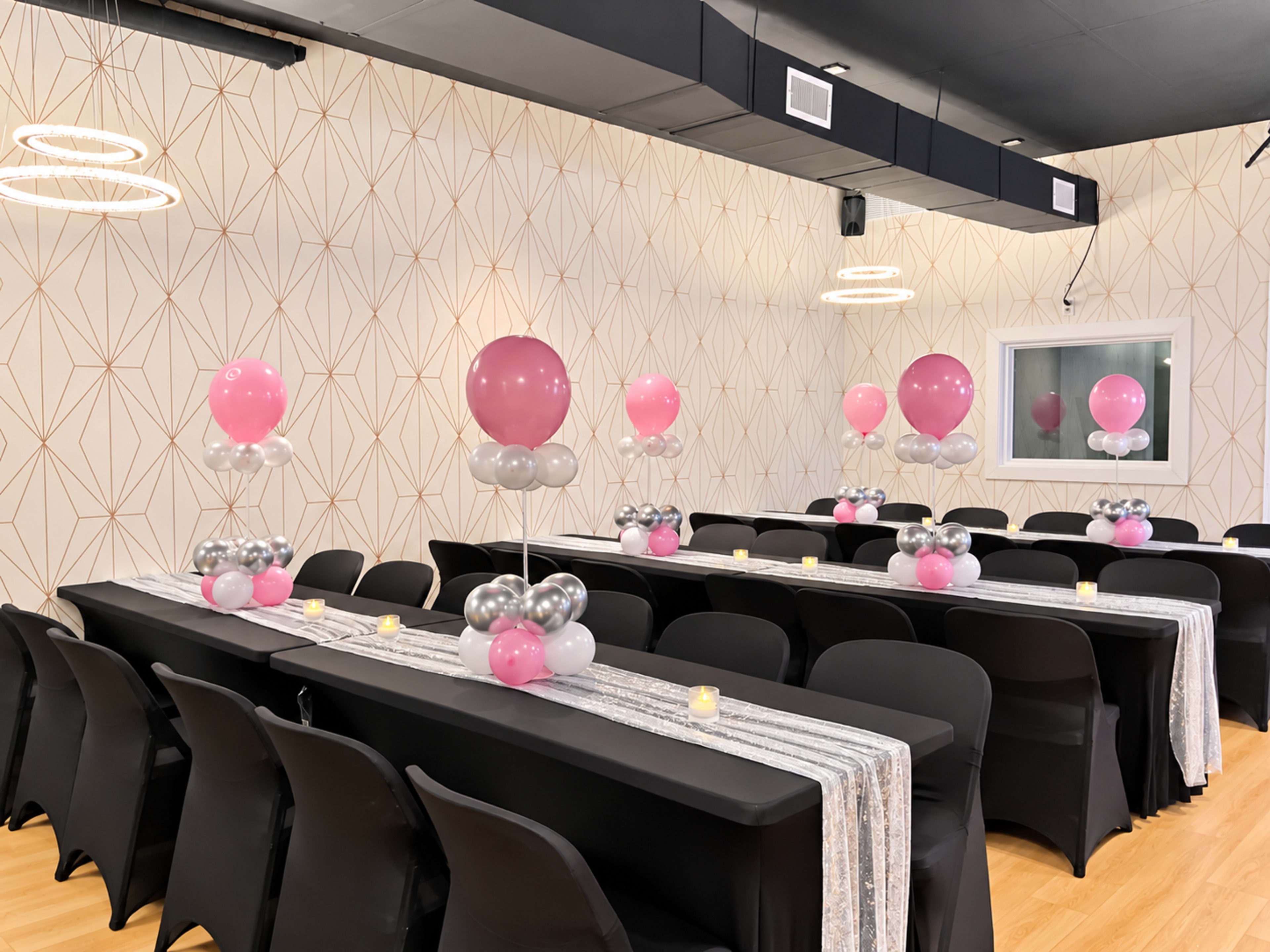 The image shows a decorated event space featuring long black tables with pink and silver balloon centerpieces and lit candles along a table runner.