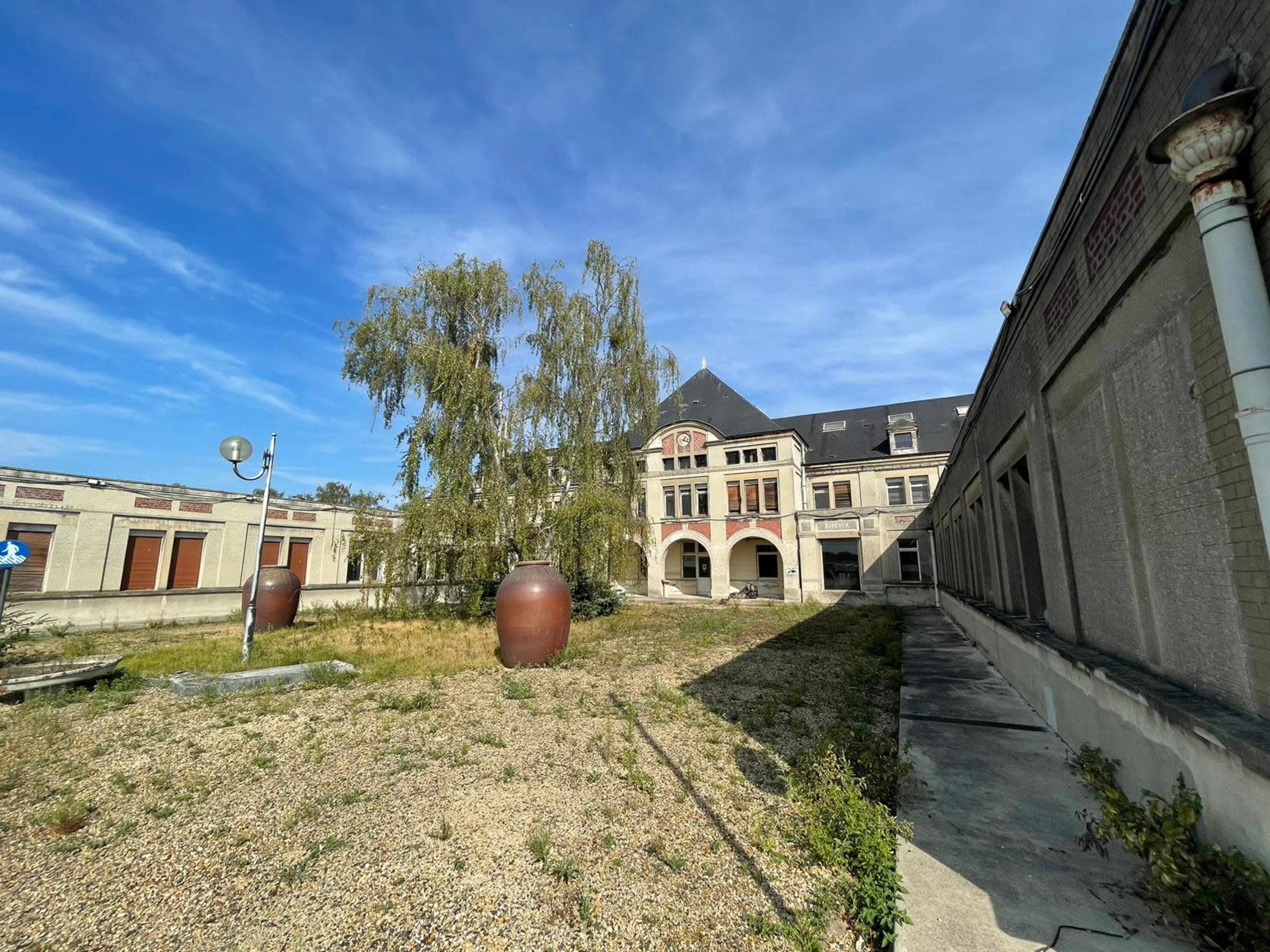 The image shows a vacant courtyard with a large building in the background, surrounded by overgrown grass and two large clay pots.