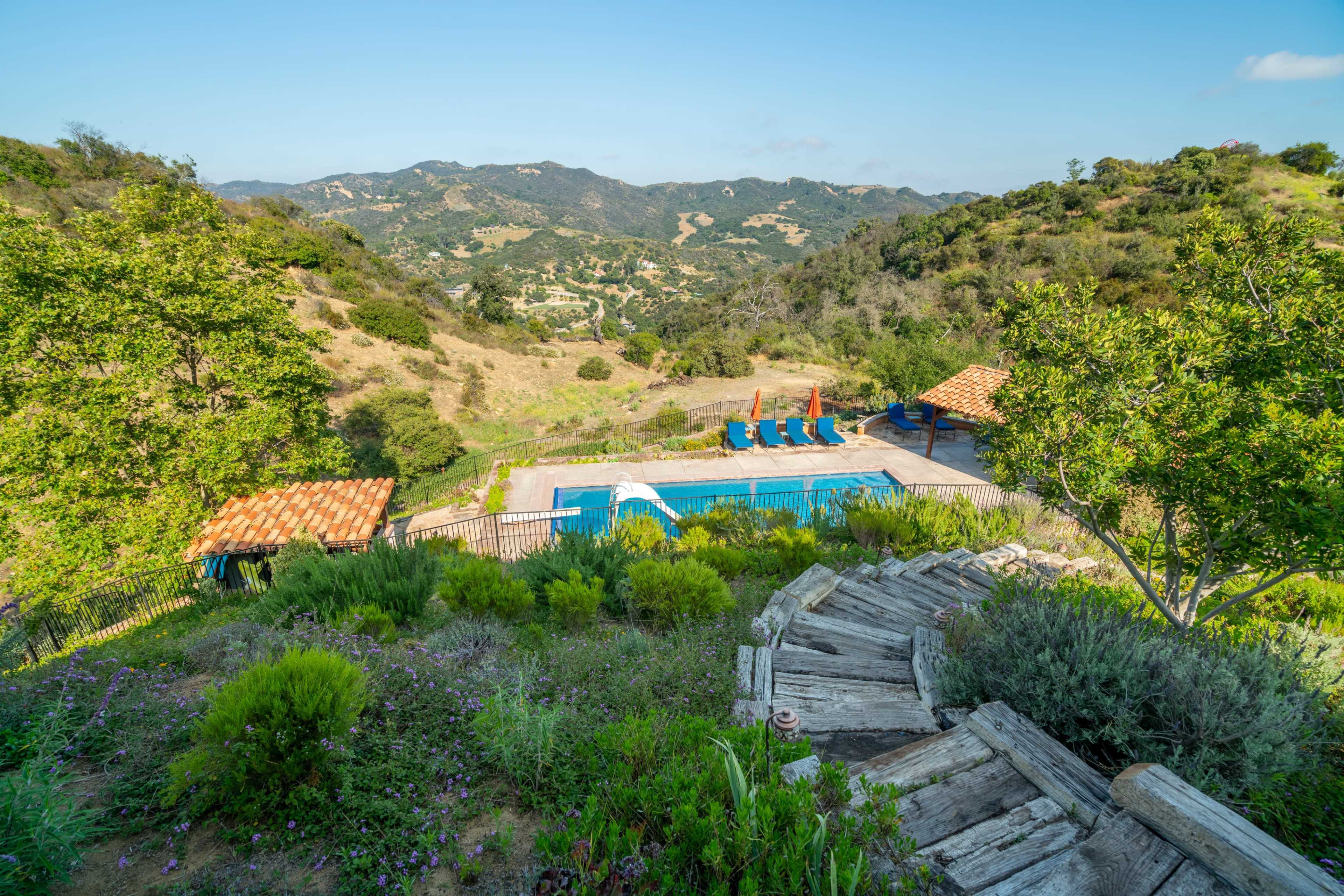 A hillside property features a pool surrounded by lounge chairs, with a view of rolling hills and trees in the background.