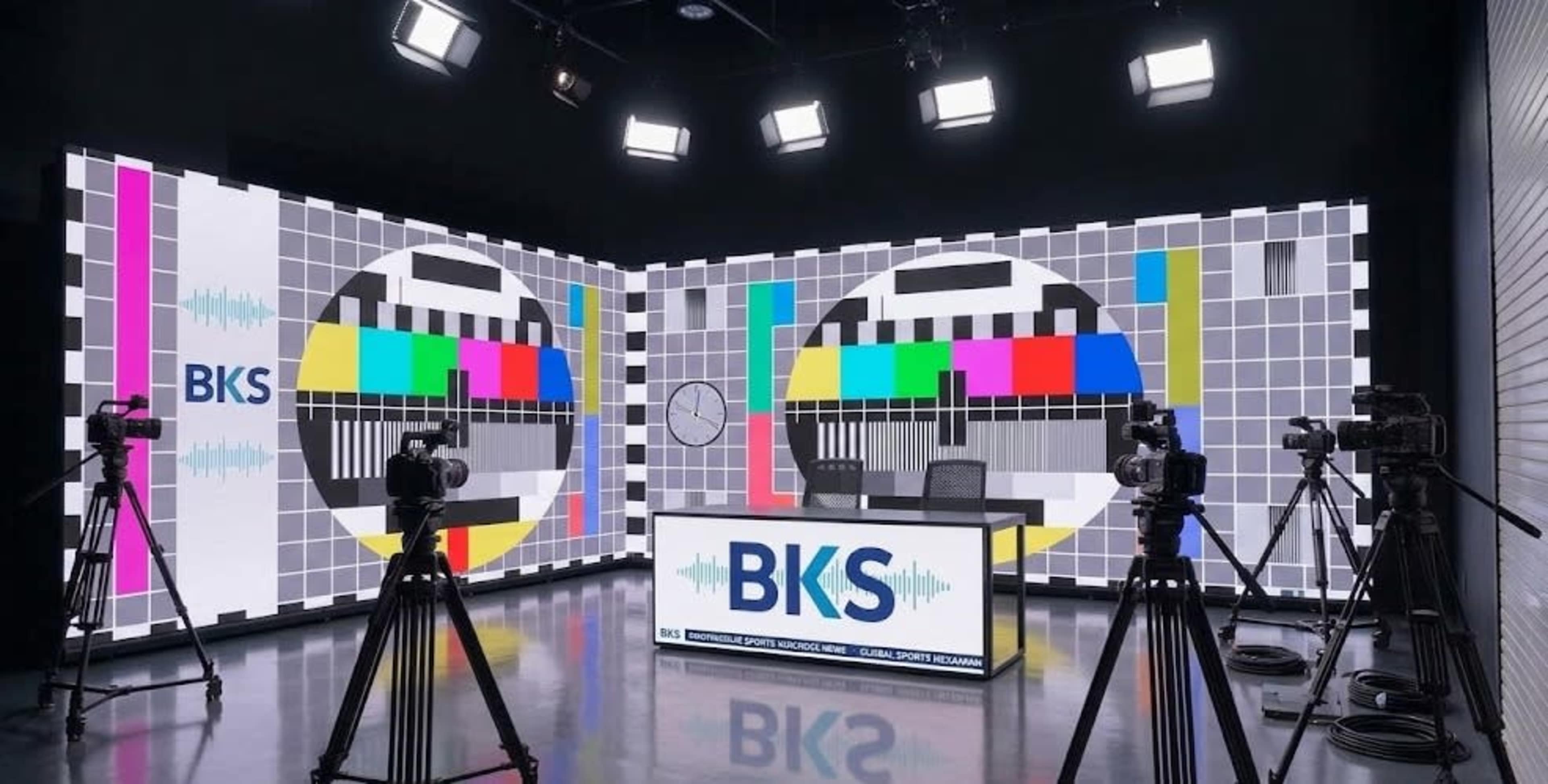 The image depicts a modern broadcasting studio with a large test pattern backdrop, camera equipment, and a centrally positioned desk featuring the logo "BKS."