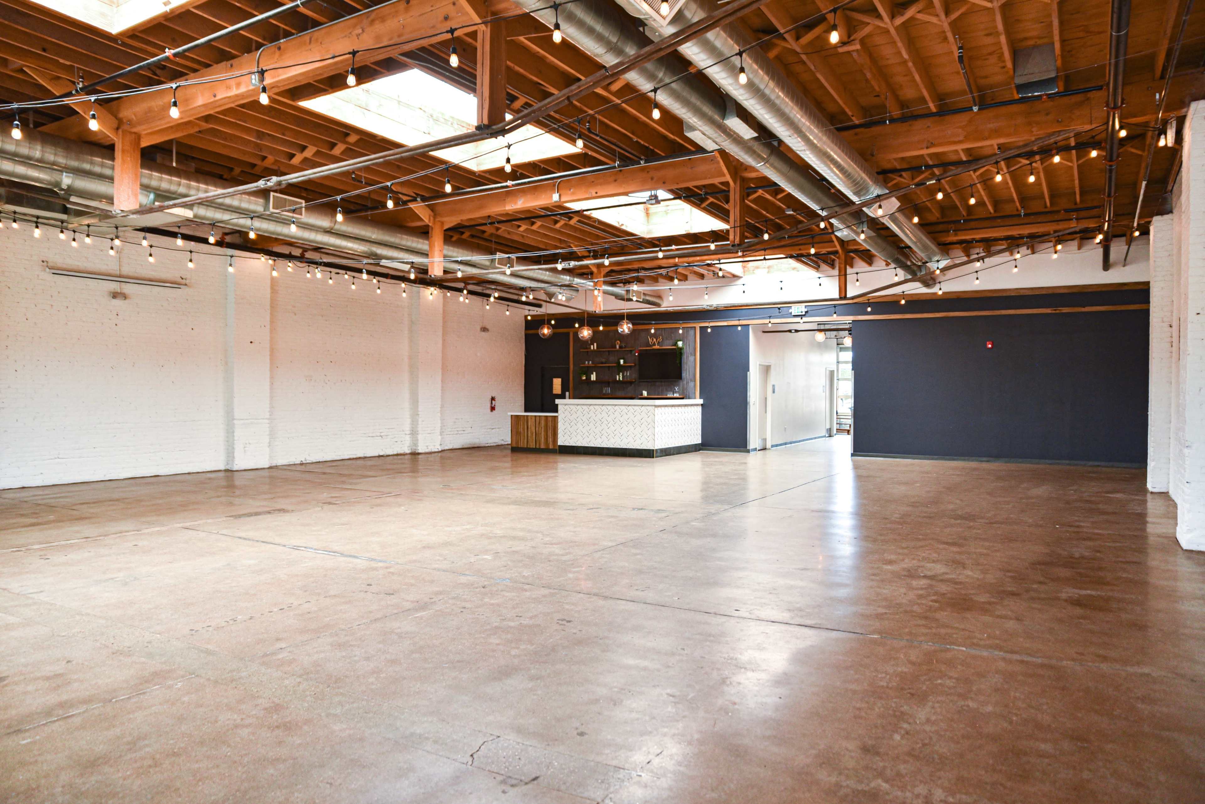 The image shows a spacious, well-lit interior with polished concrete flooring, exposed wooden beams, and metal ductwork, featuring a bar area and minimal decor.
