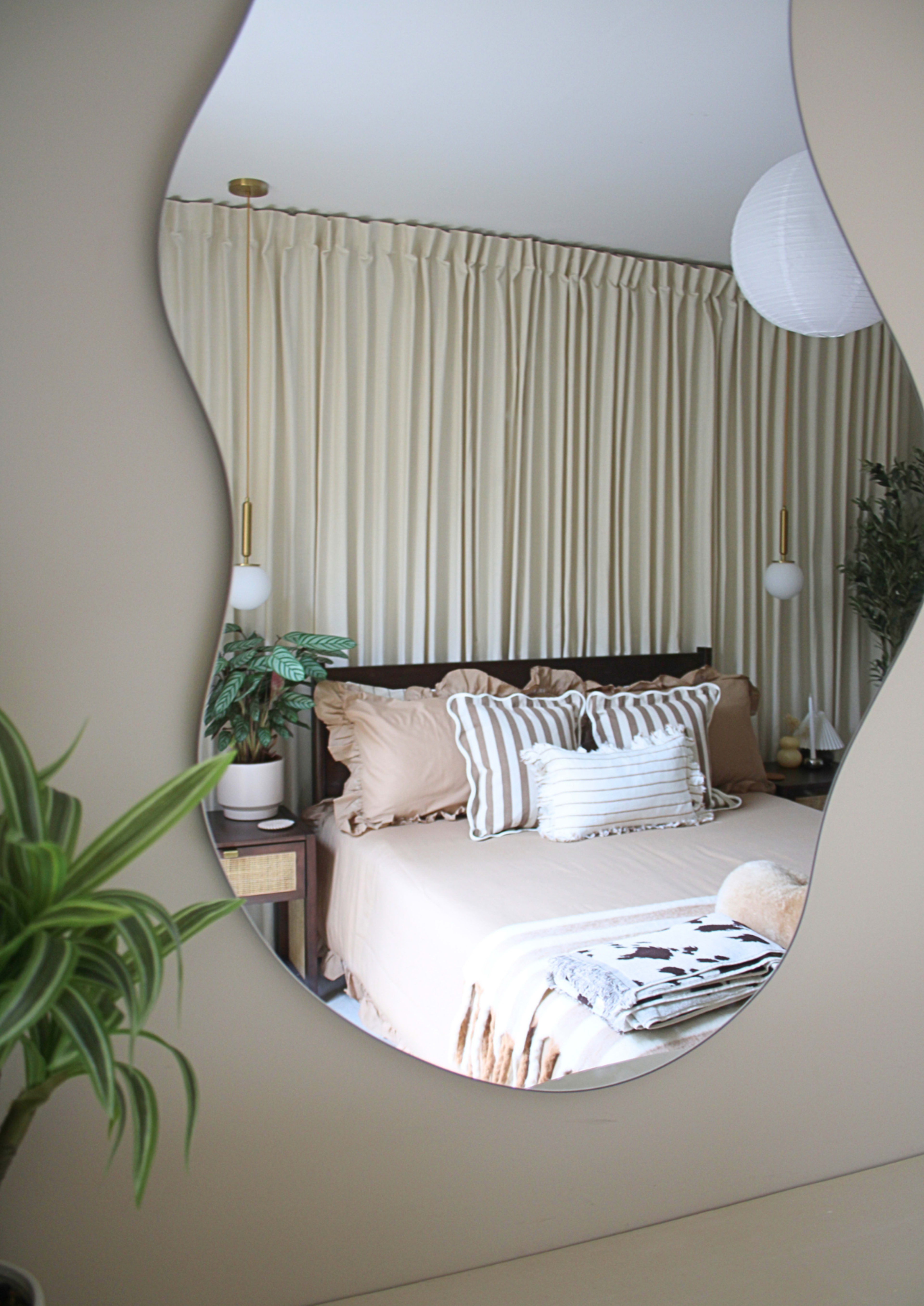The image shows a softly illuminated bedroom with a reflective, wavy mirror, a bed with neutral-toned bedding, and a potted plant in the foreground.