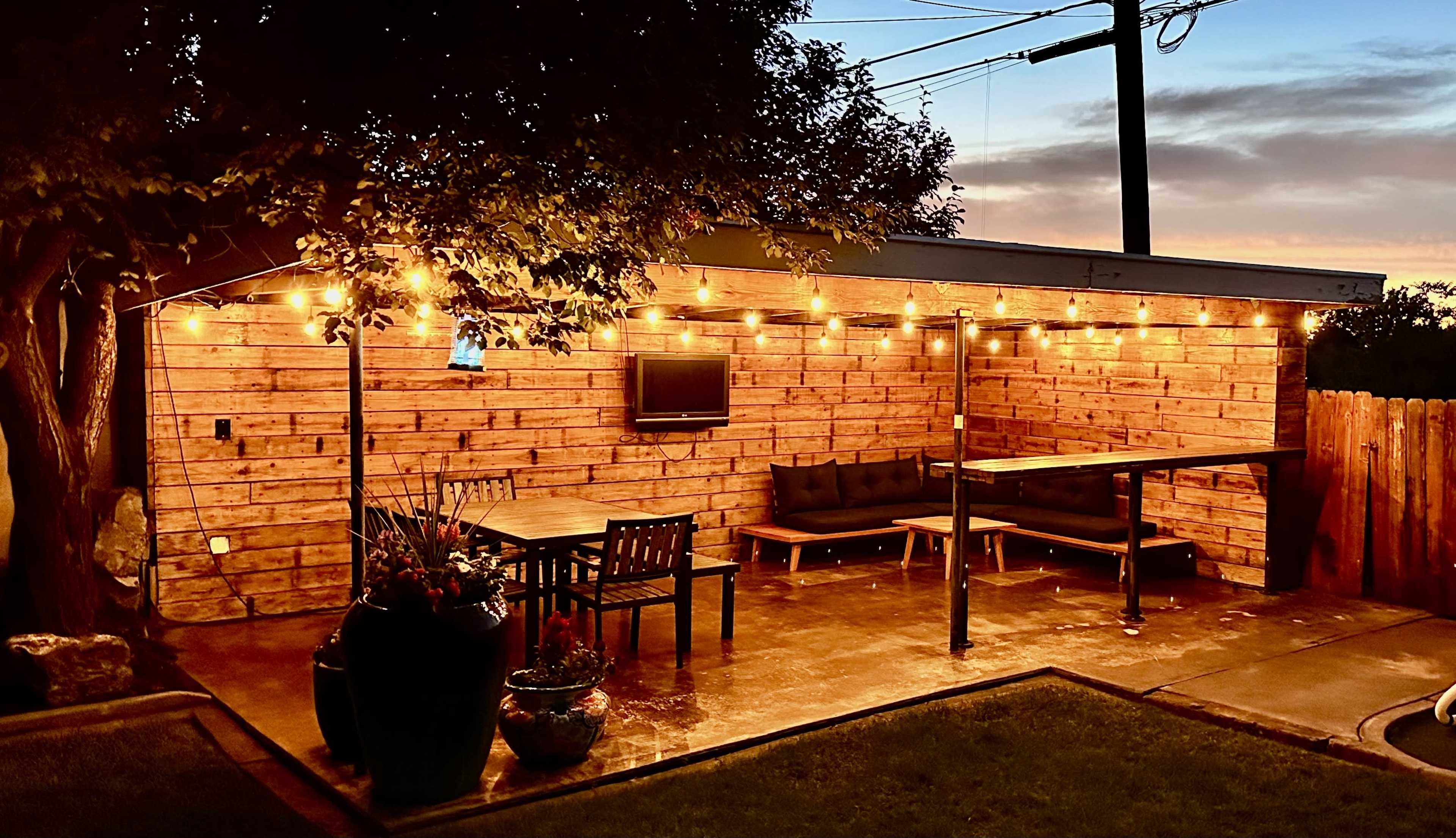 The image shows a lit outdoor patio area featuring wooden walls, a dining table with chairs, and a television mounted on the wall, surrounded by plants at dusk.