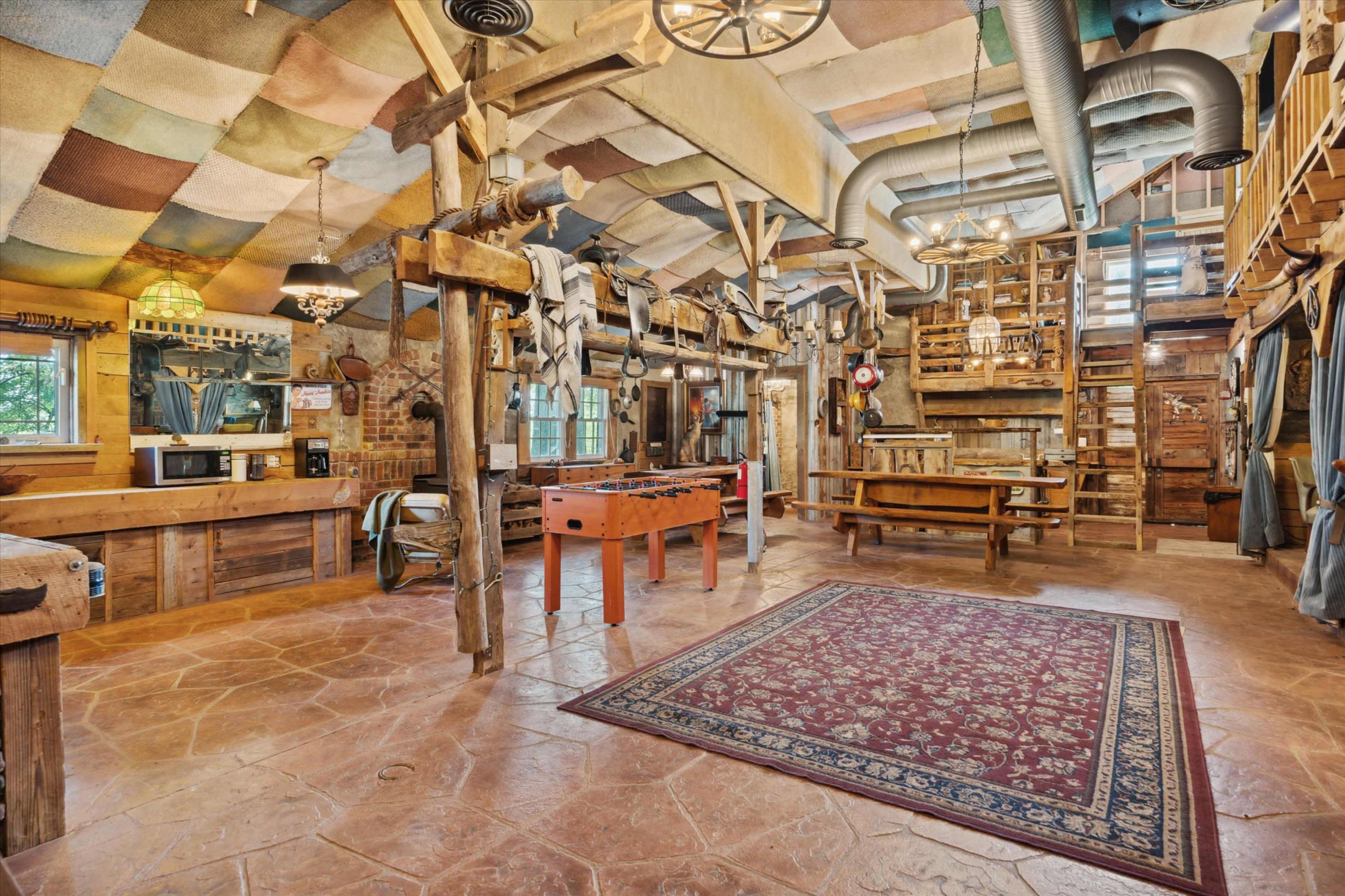 The image shows a rustic interior space featuring wooden beams, a foosball table, a large dining table, and a patterned ceiling.