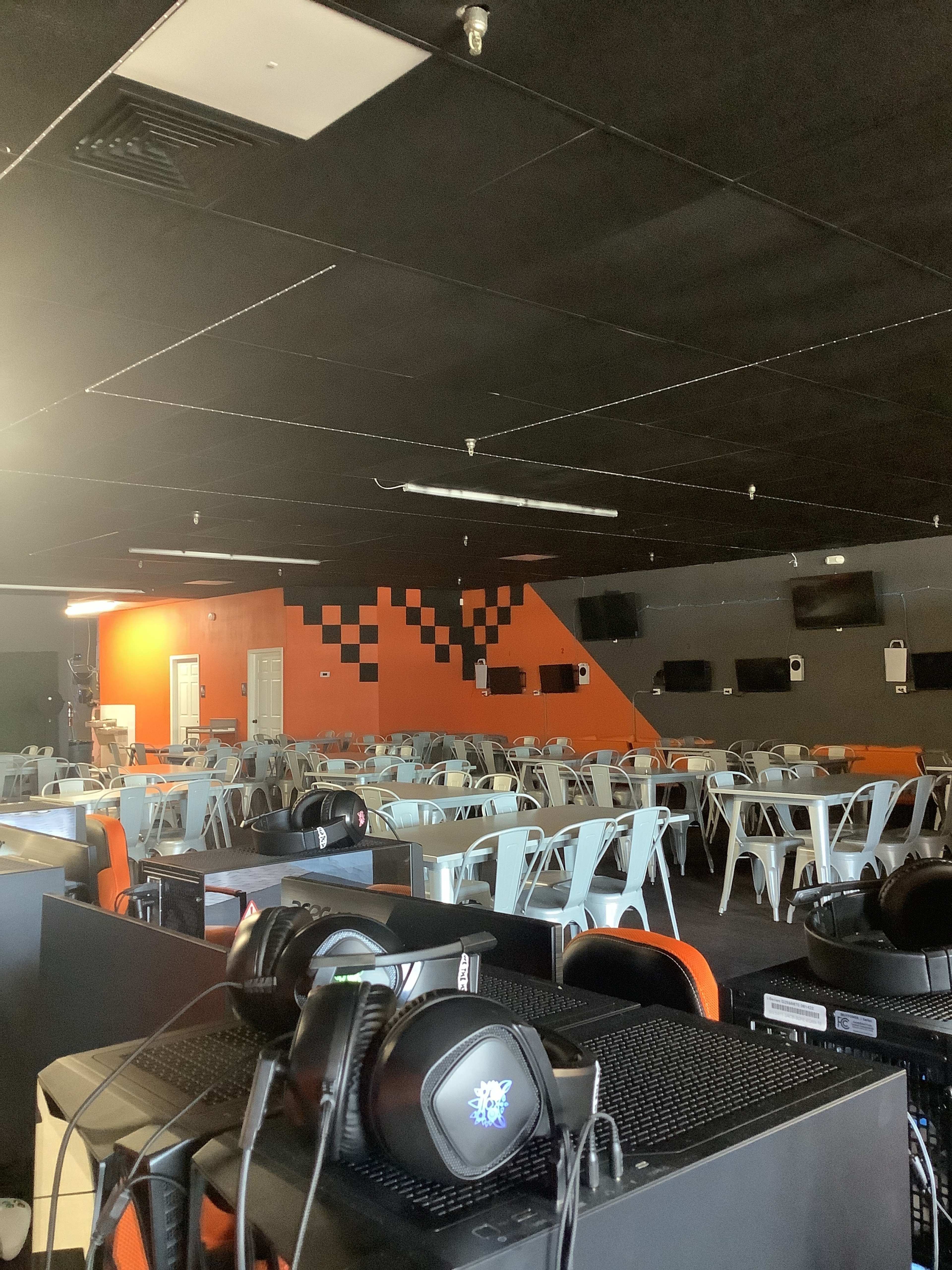 The image shows a spacious gaming lounge with tables, chairs, and several computer setups against a backdrop of orange and black walls.