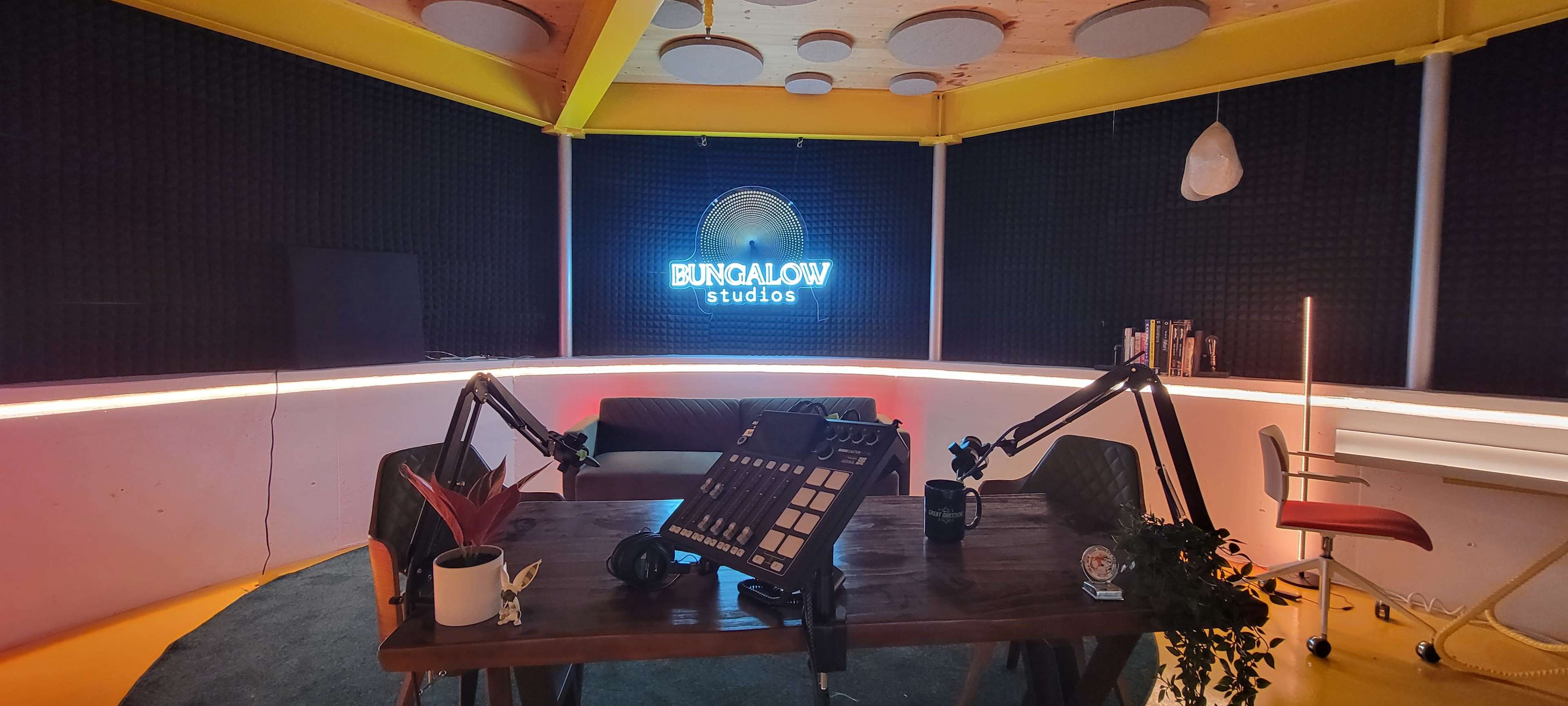 The image shows a recording studio with a neon sign that reads "Bungalow Studios," featuring a central table, microphone stands, and seating arrangements against a backdrop of soundproofing panels.