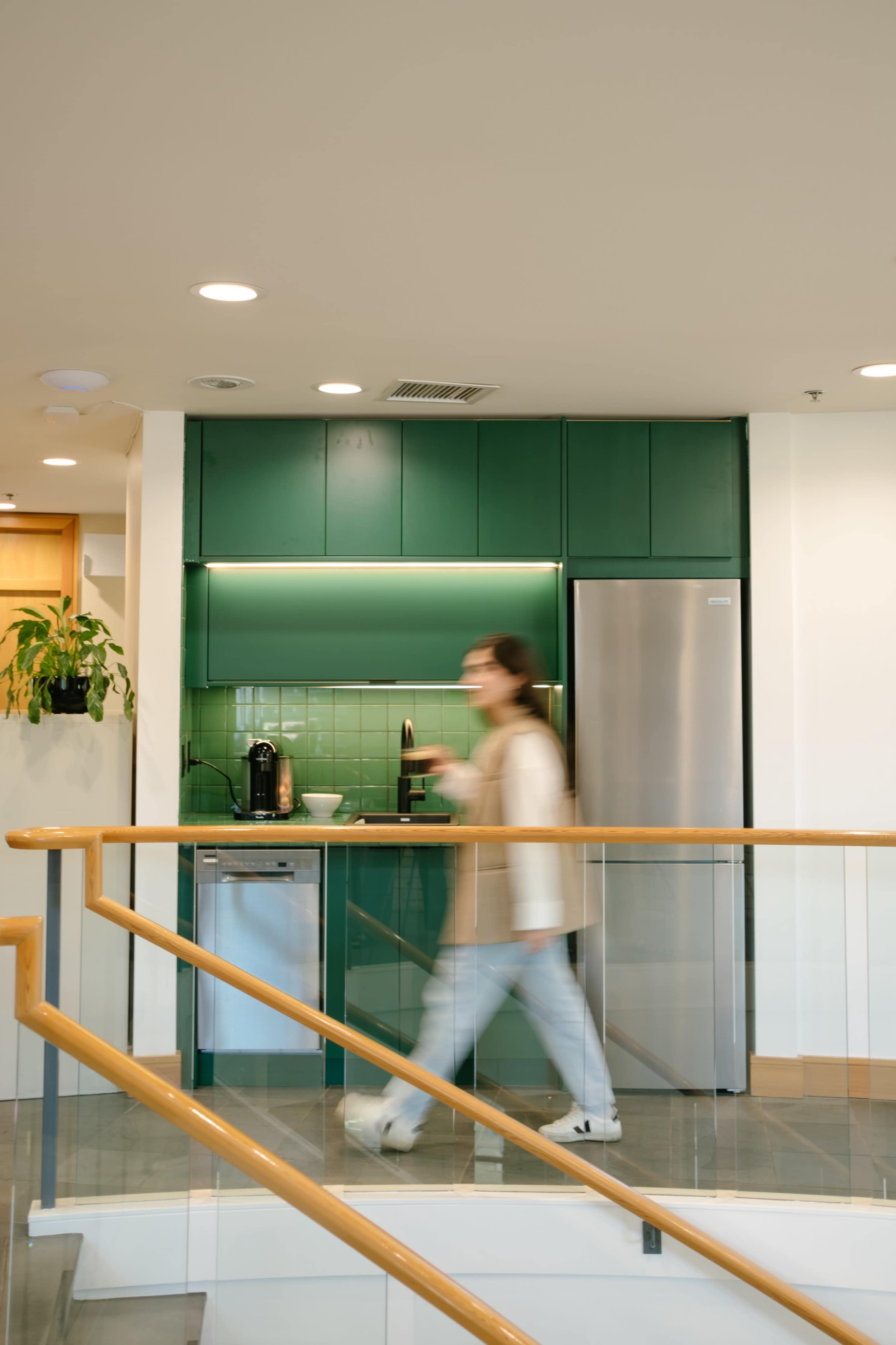 A person walks past a modern kitchen area with green tiles, a refrigerator, and a coffee maker.