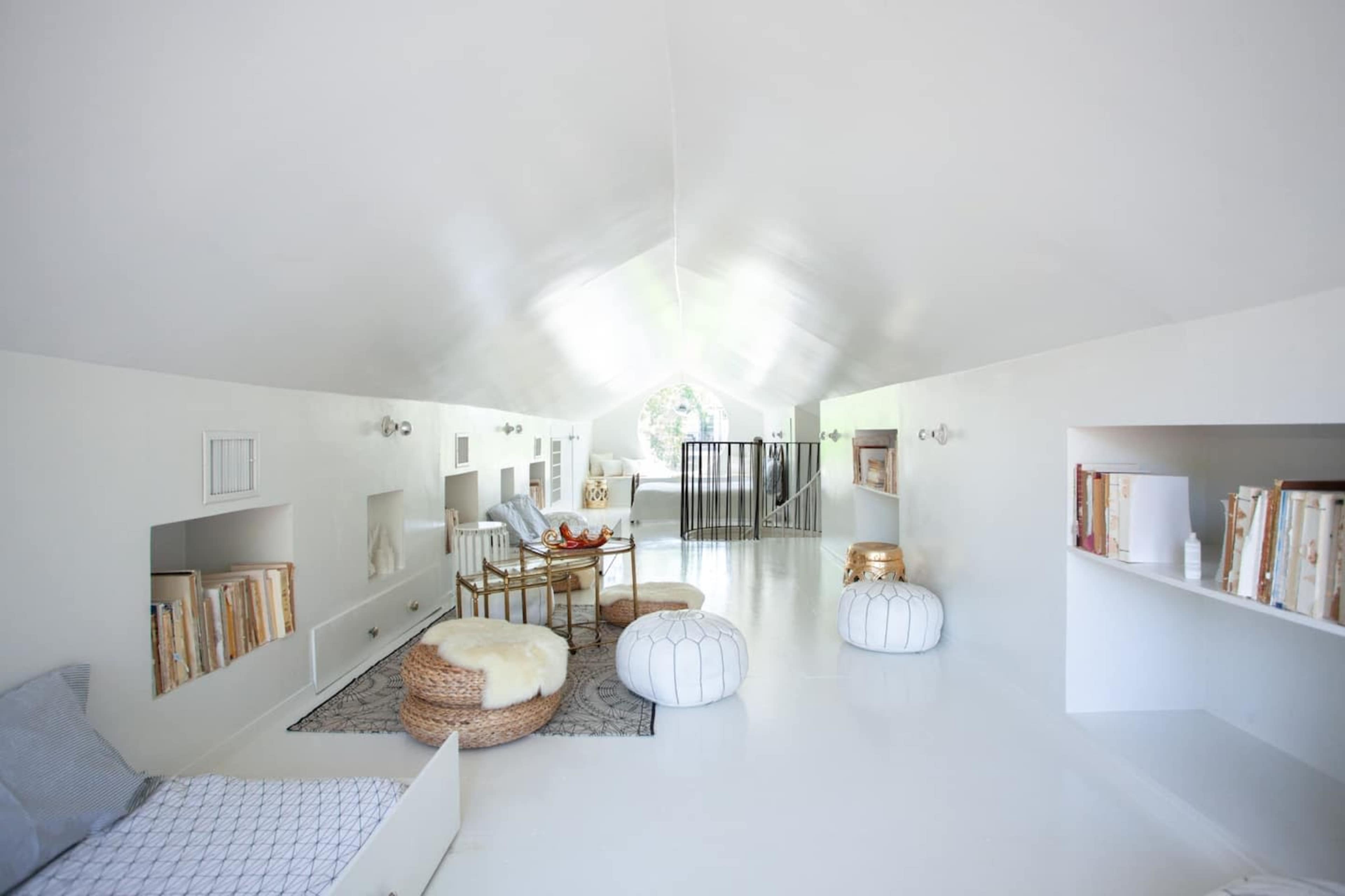 A minimalist, brightly lit interior space with white walls, a wooden ceiling, and various seating options, including poufs and a woven rug.