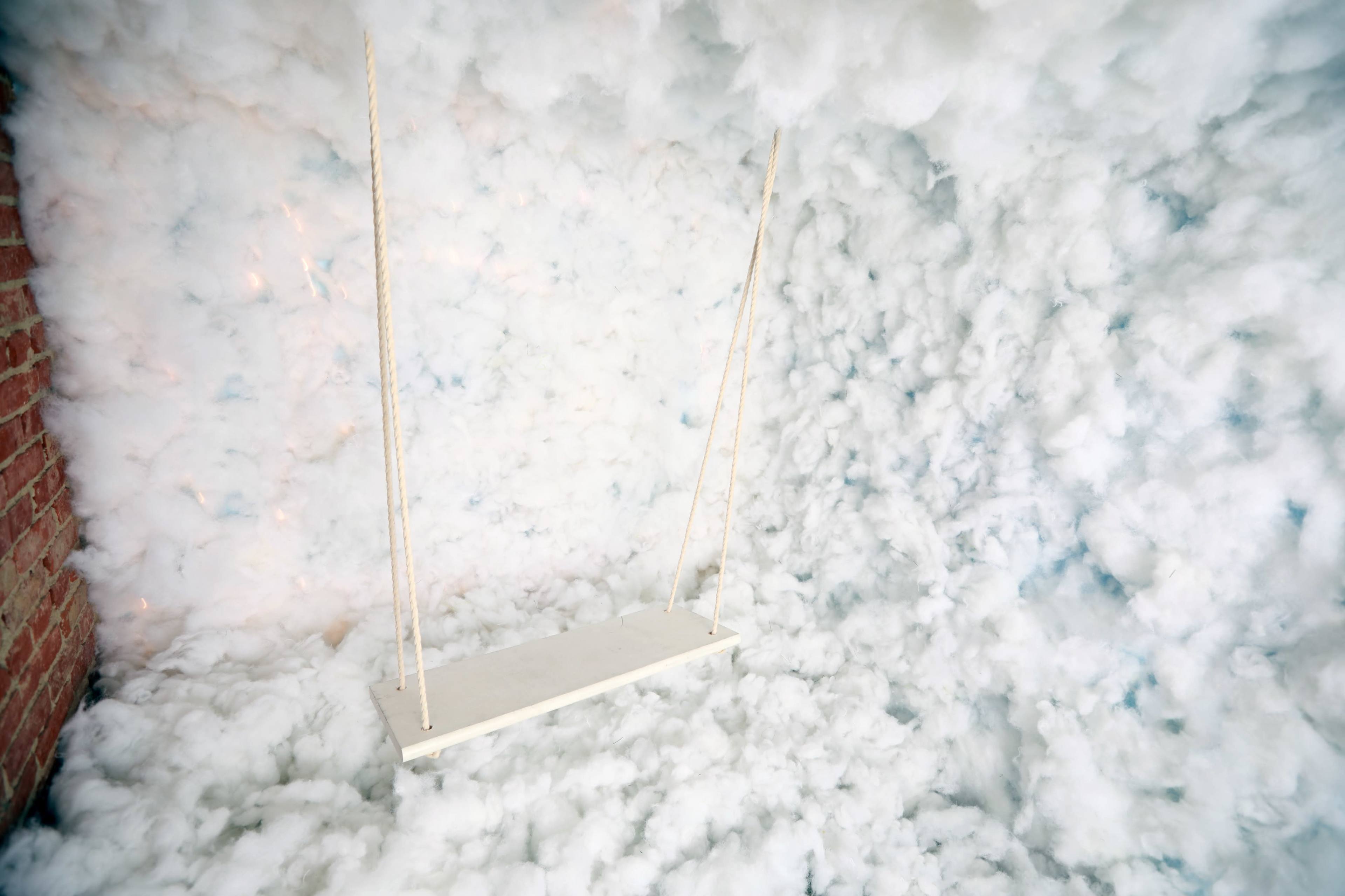 A white swing hangs from ropes amid a backdrop of fluffy white material resembling clouds.