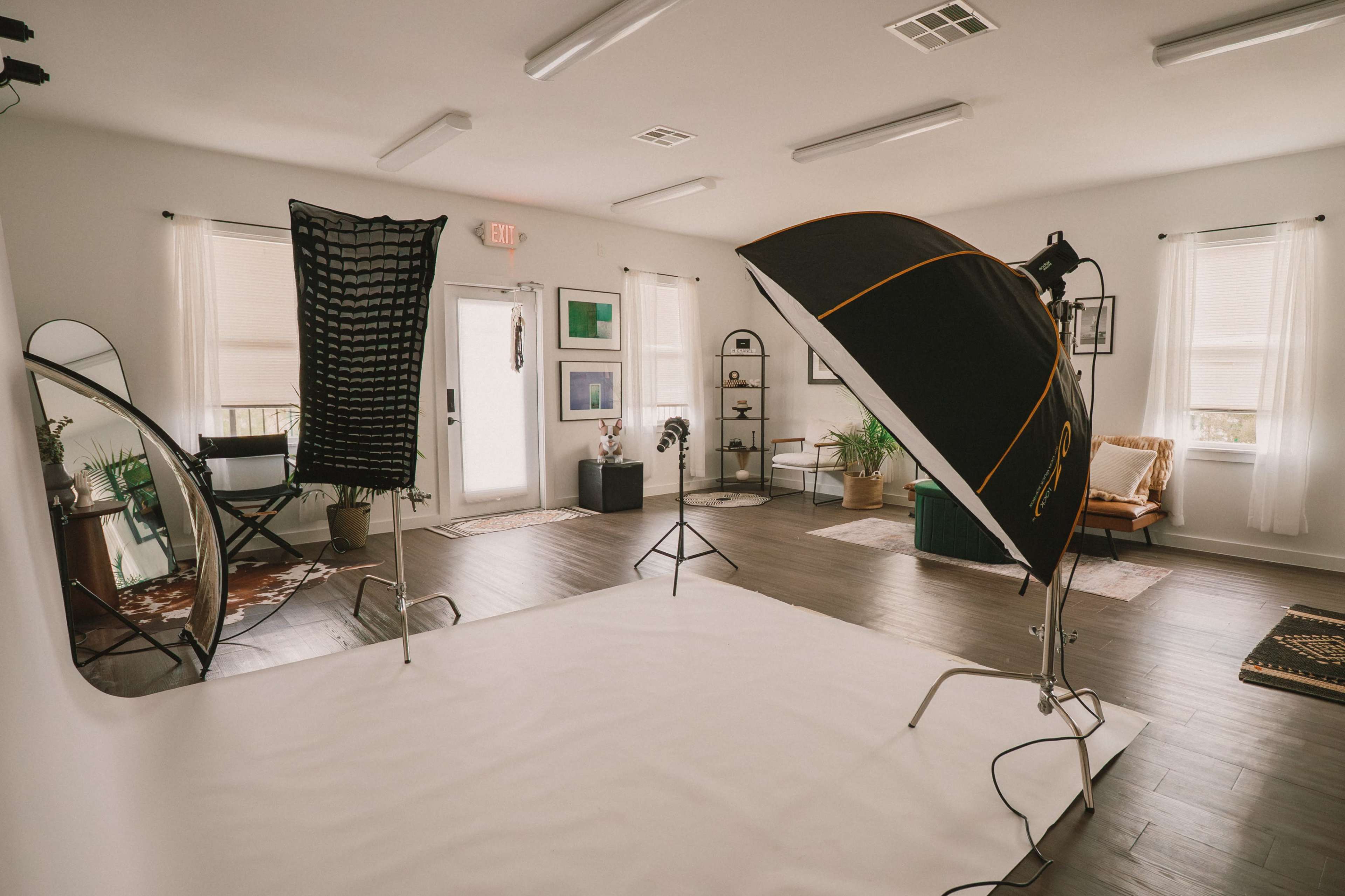 The image shows a well-lit photography studio featuring a seamless white backdrop, various lighting equipment, and a cozy seating area with decorative elements.