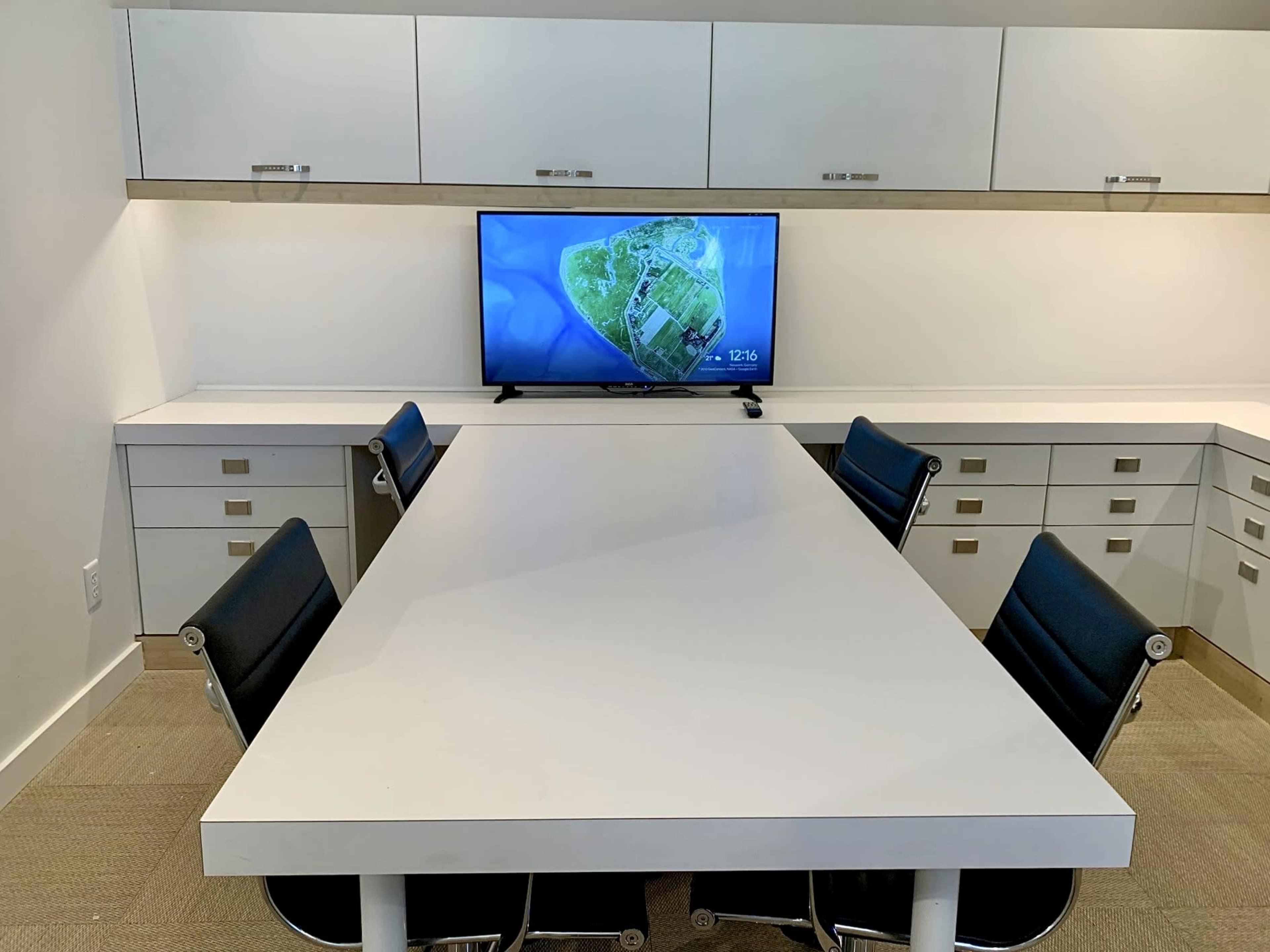 A conference room features a large white table with black chairs and a television mounted on the wall displaying an aerial view of an island.