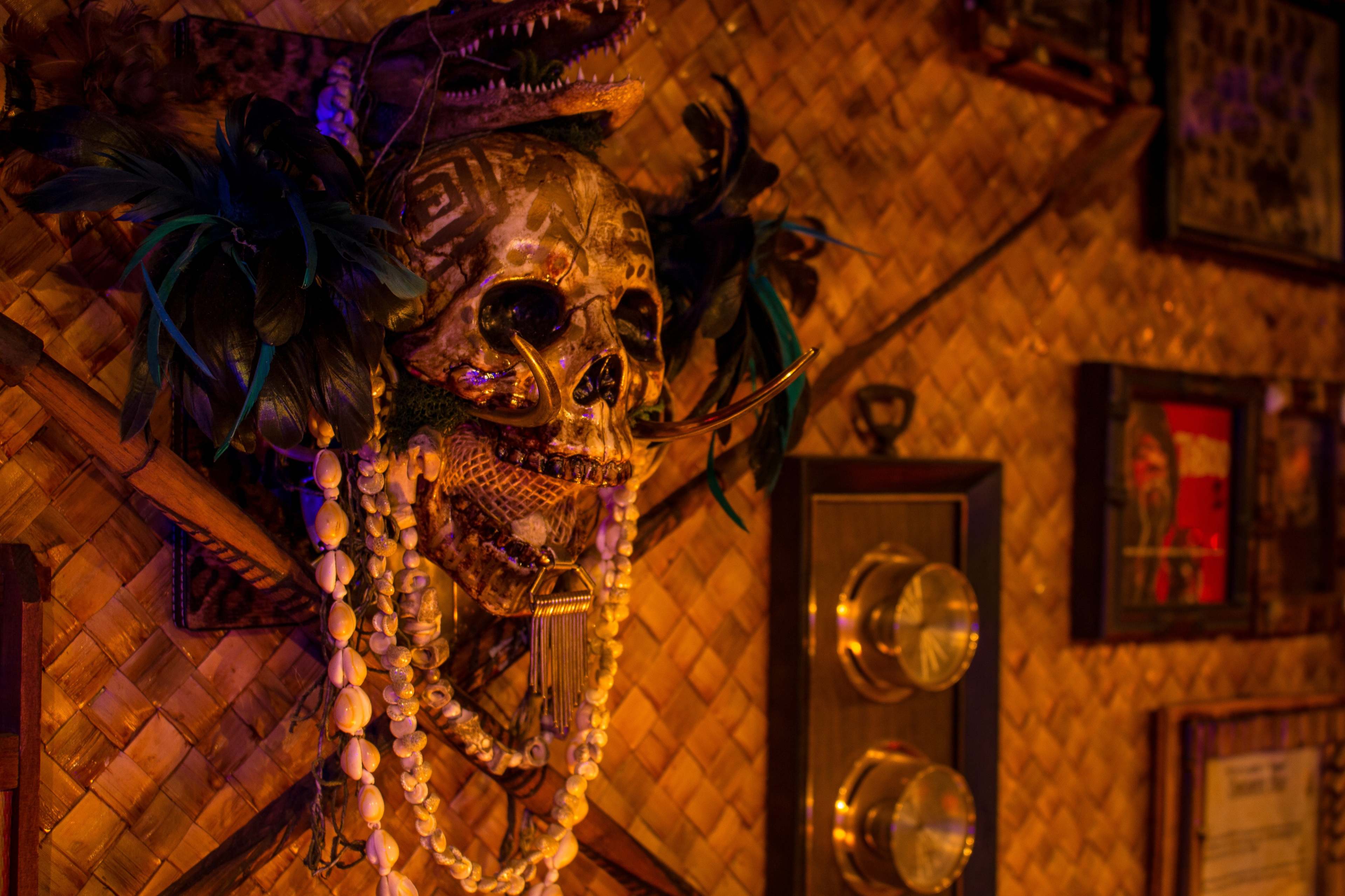 A decorative skull mounted on a woven bamboo wall, adorned with feathers and beads, alongside framed pictures and vintage hardware.