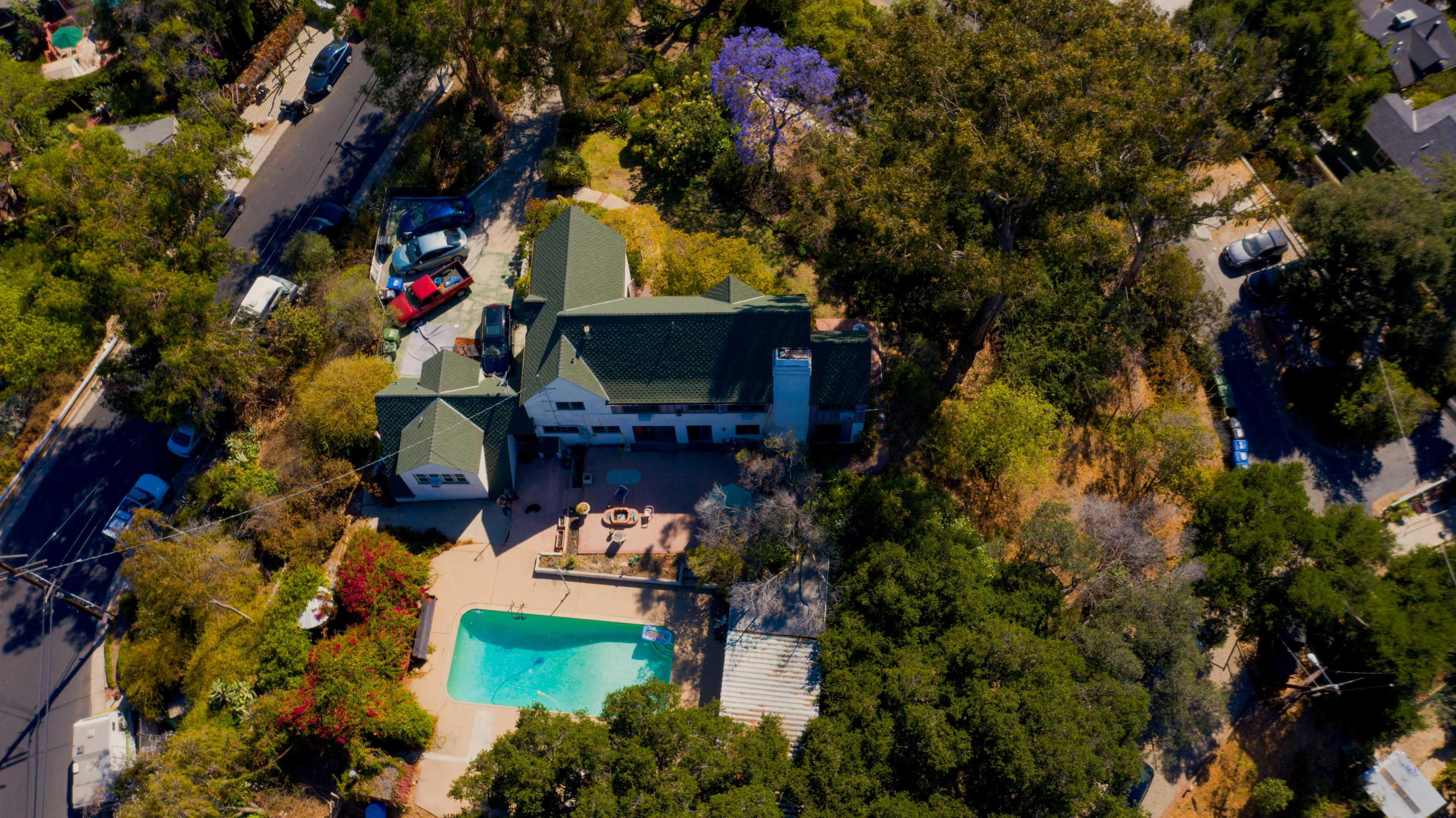 An aerial view of a house surrounded by greenery, featuring a swimming pool and several parked cars nearby.