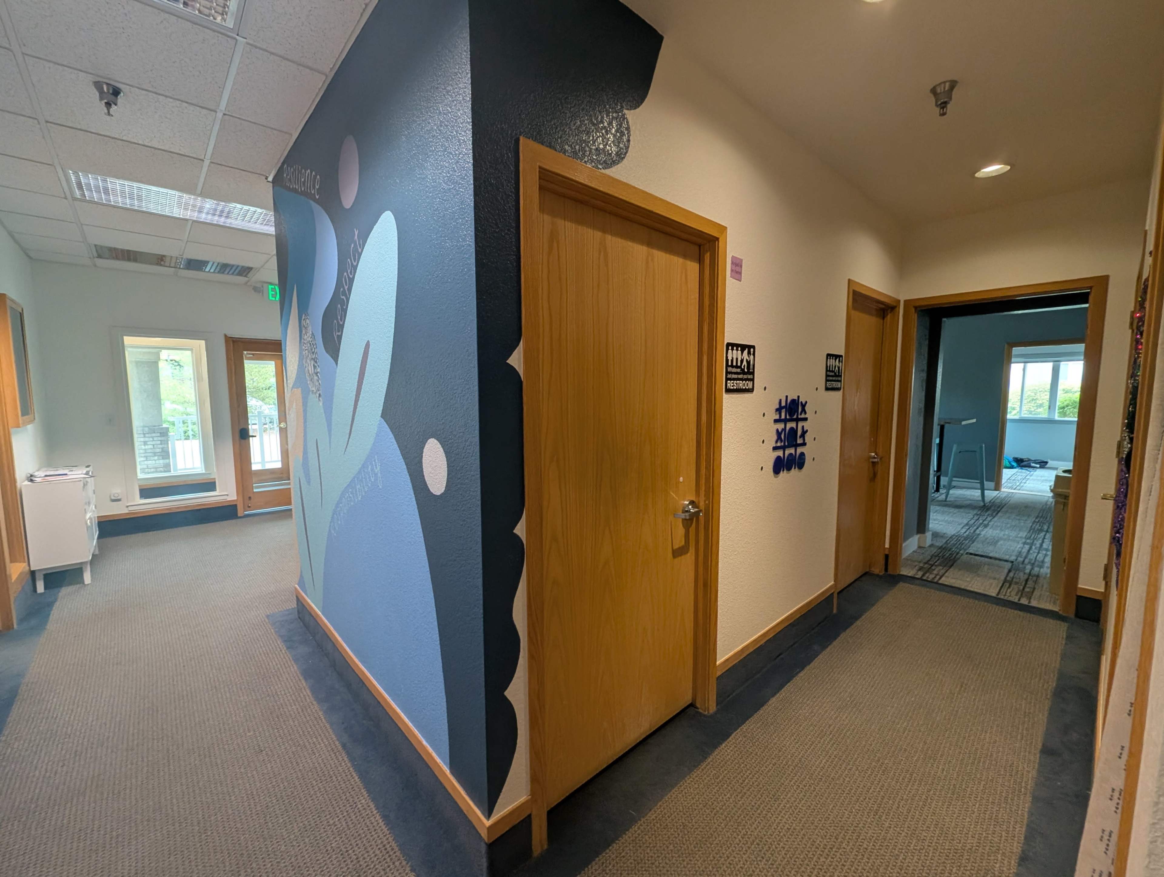 The image depicts a hallway featuring a blue and white mural on one wall, with wooden doors leading to adjacent rooms on either side.