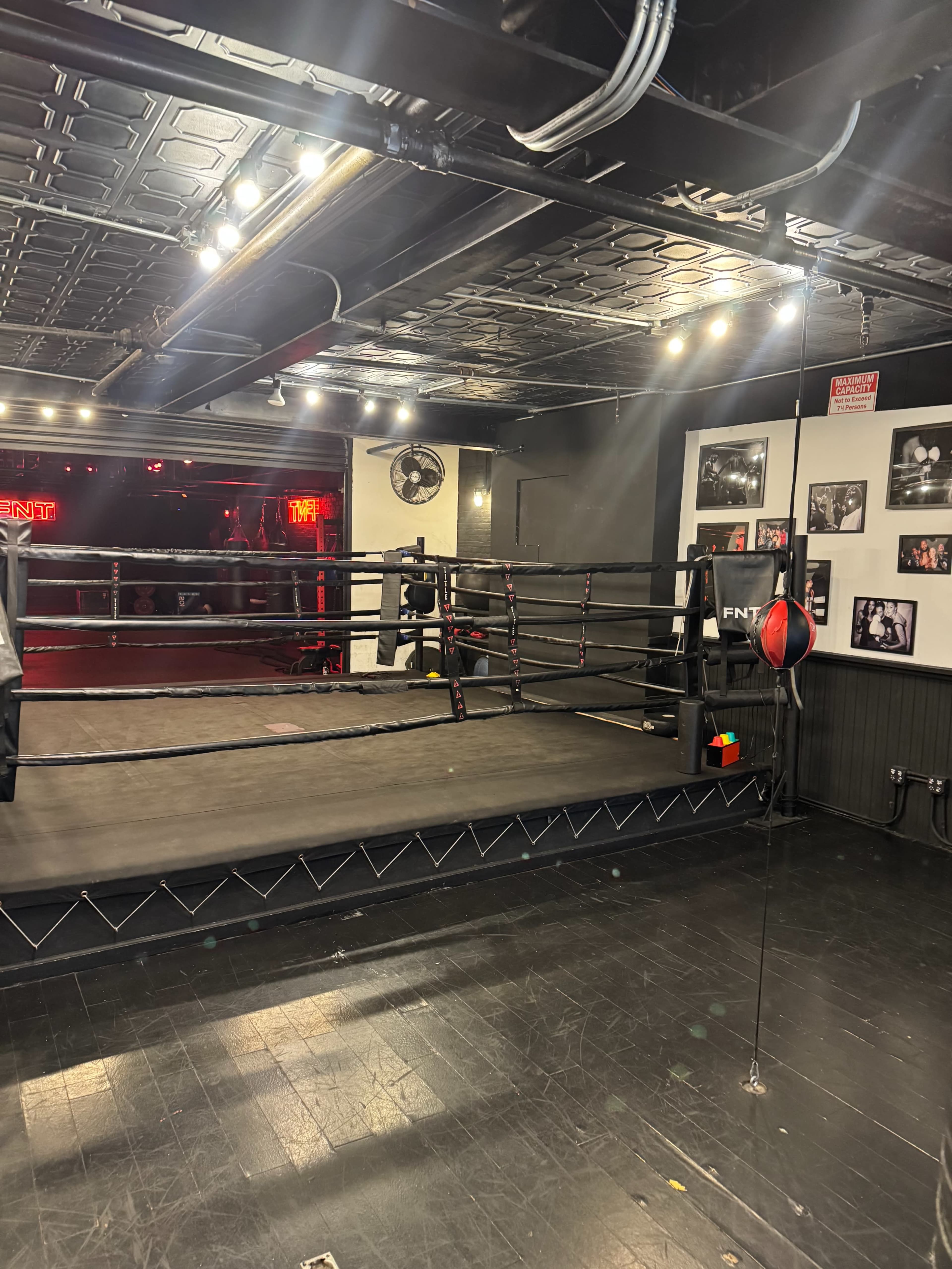 The image shows an empty boxing ring in a dimly lit gym, with black flooring and framed photographs on the walls.