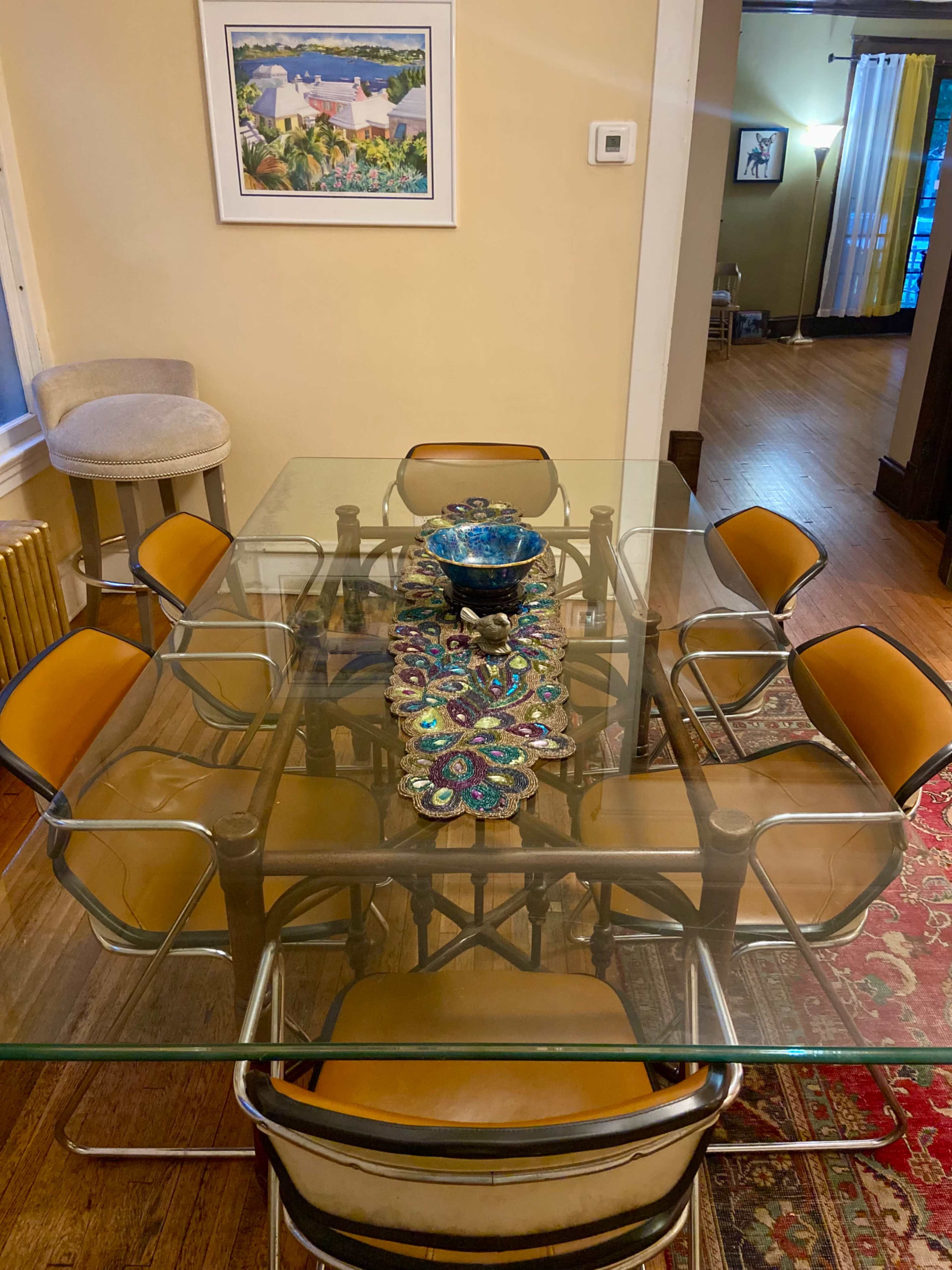 A glass-top dining table with a colorful centerpiece is surrounded by six chairs in a well-lit room.