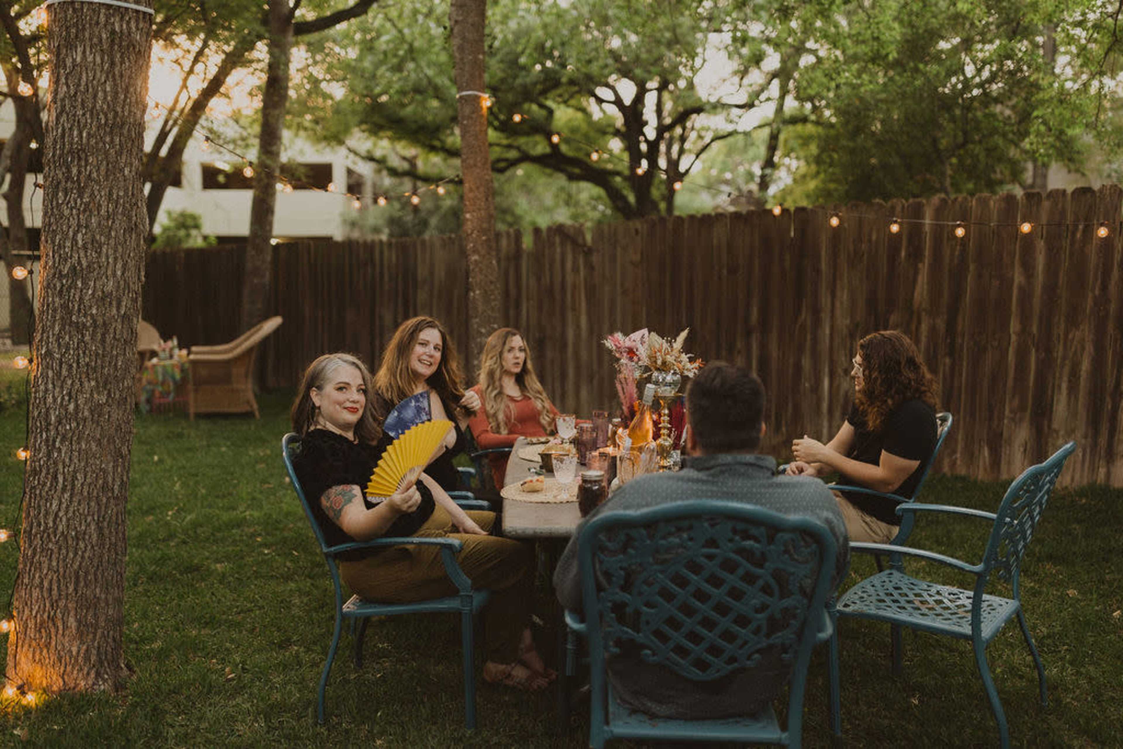 A group of five people sits around a table in a backyard, enjoying drinks and conversation amidst trees and string lights.