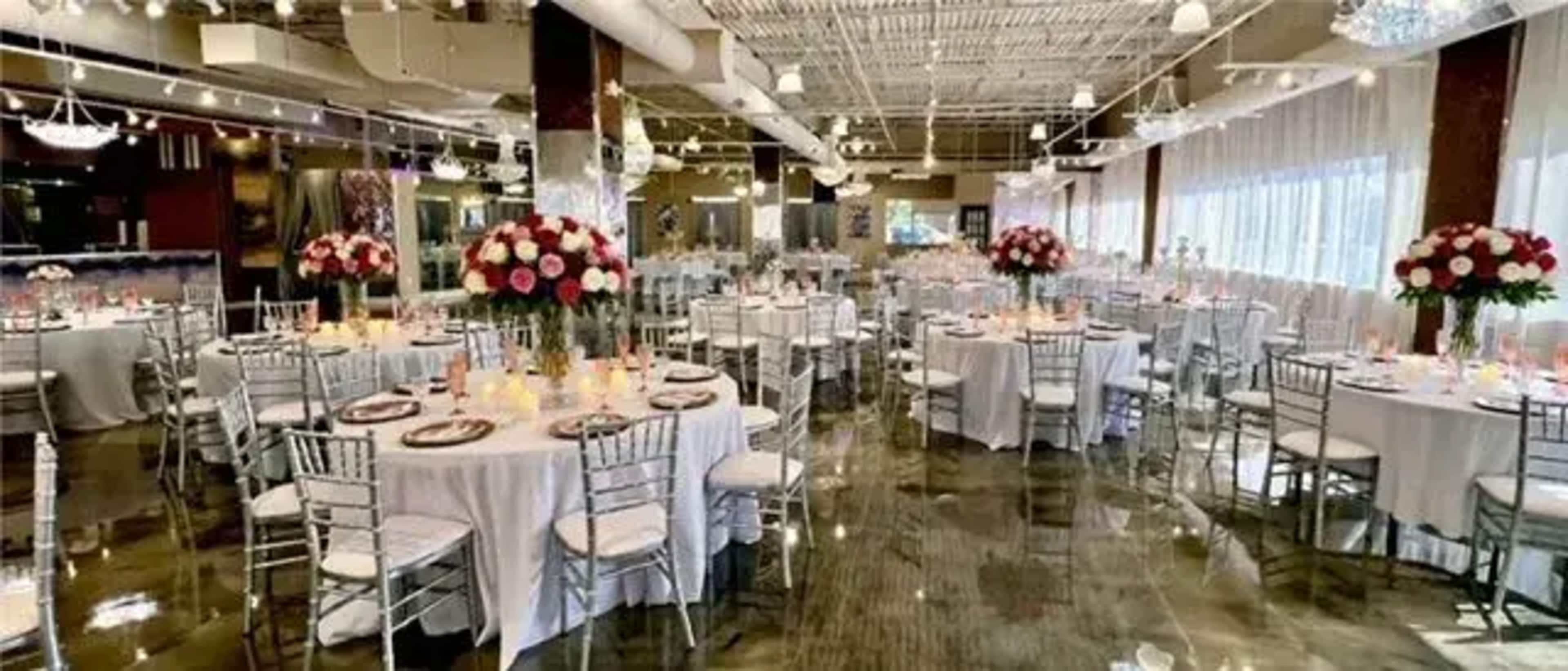 The image shows a banquet hall set for a formal event, featuring circular tables adorned with white tablecloths, elegant centerpieces of roses, and ambient lighting.