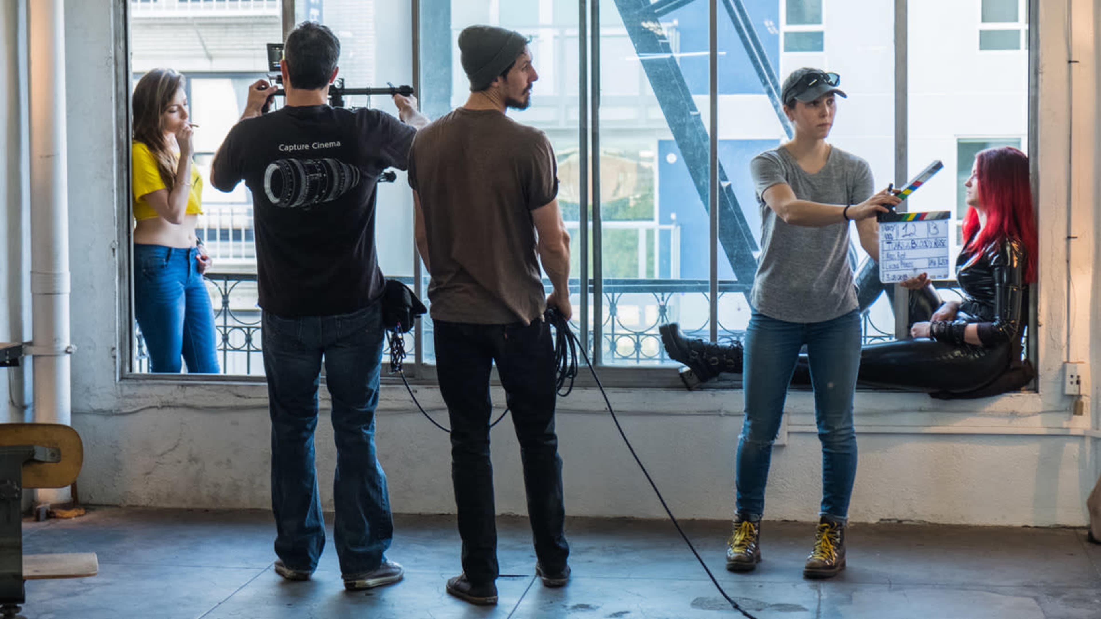A film crew sets up inside a bright room with large windows, while a woman holds a clapperboard and two people converse in the background.