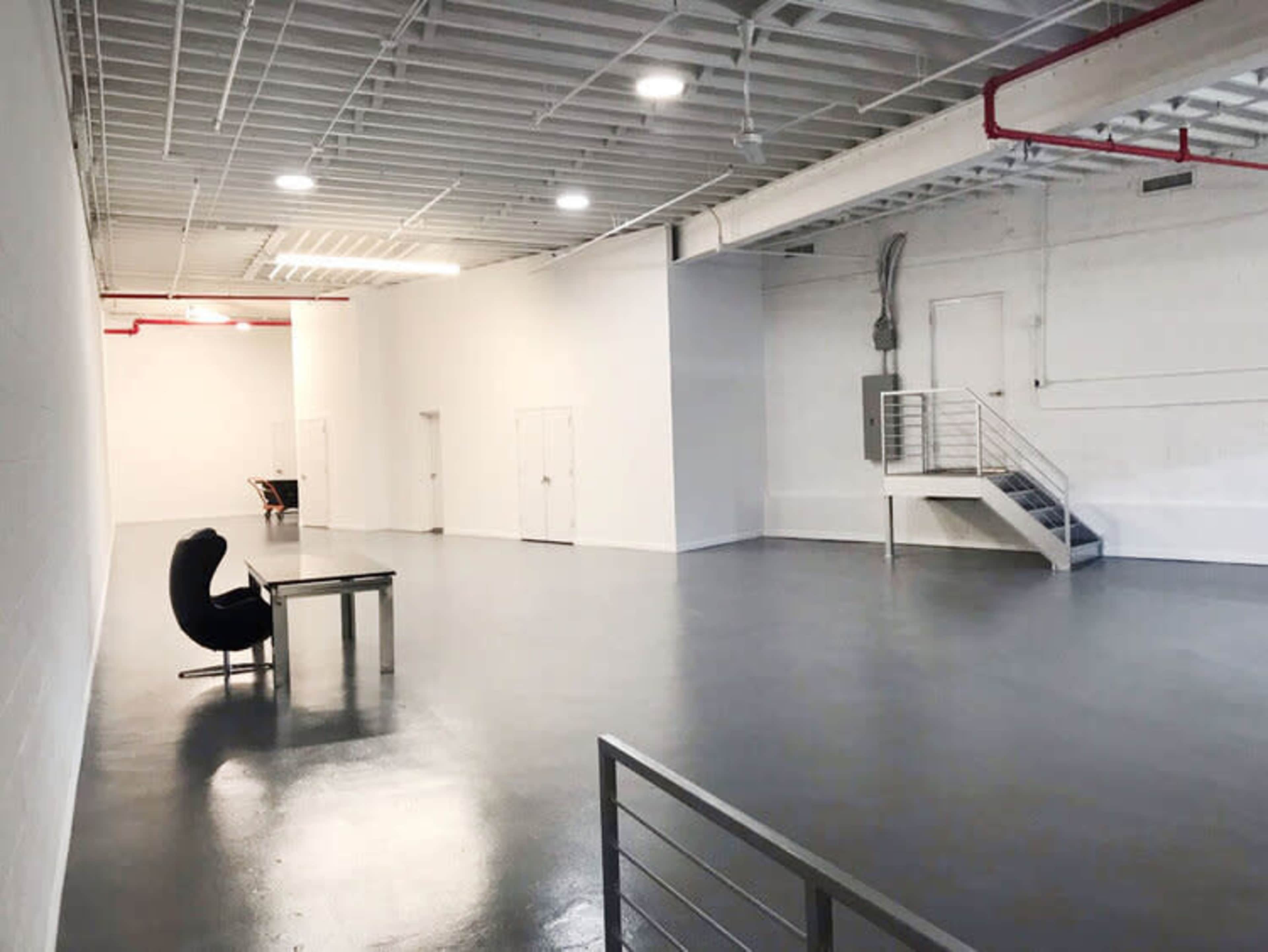 A large, empty industrial room with white walls, a gray floor, a small table, a black chair, and a staircase in the background.