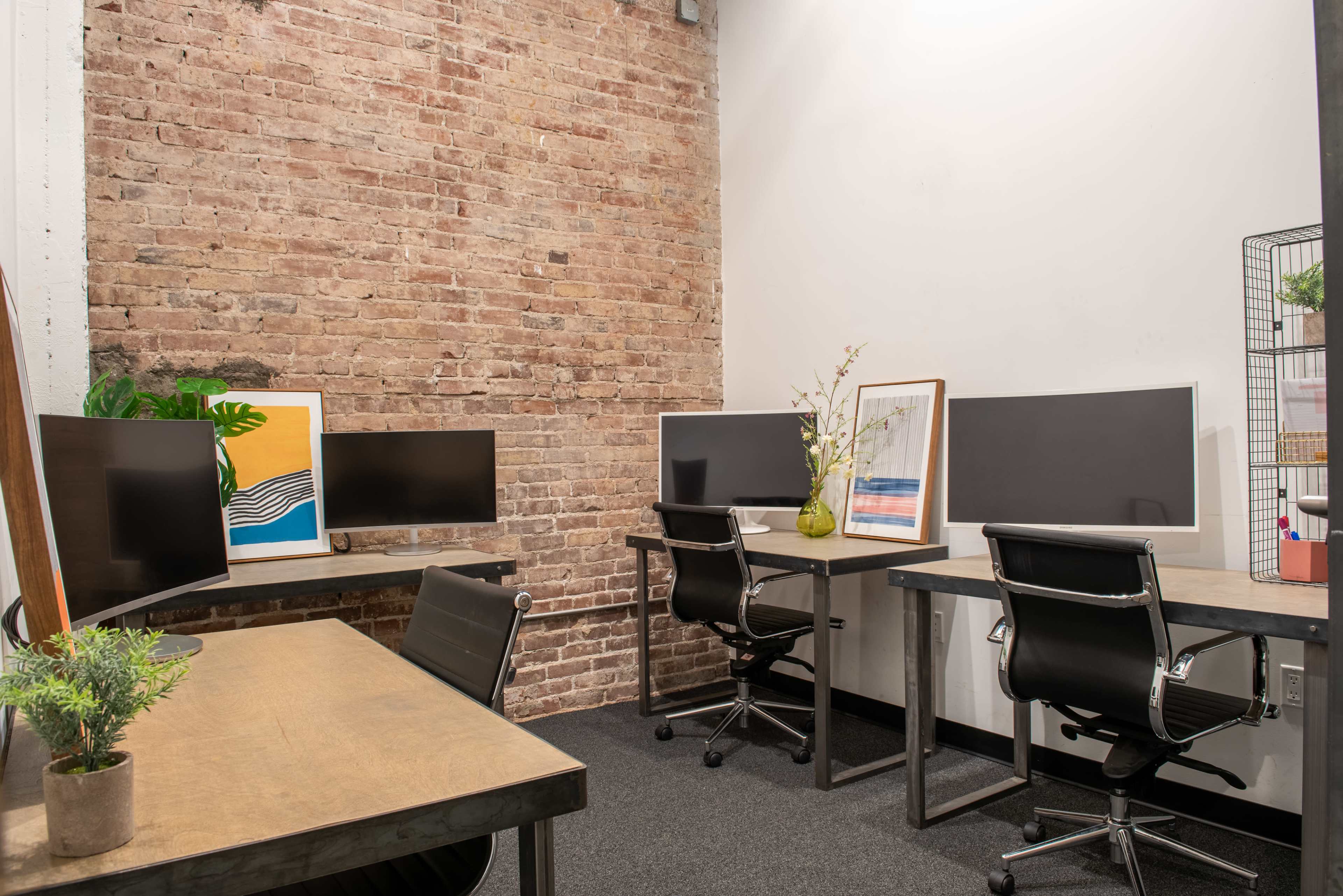 The image shows a modern office workspace with four black computer monitors arranged at two desks against a brick wall.