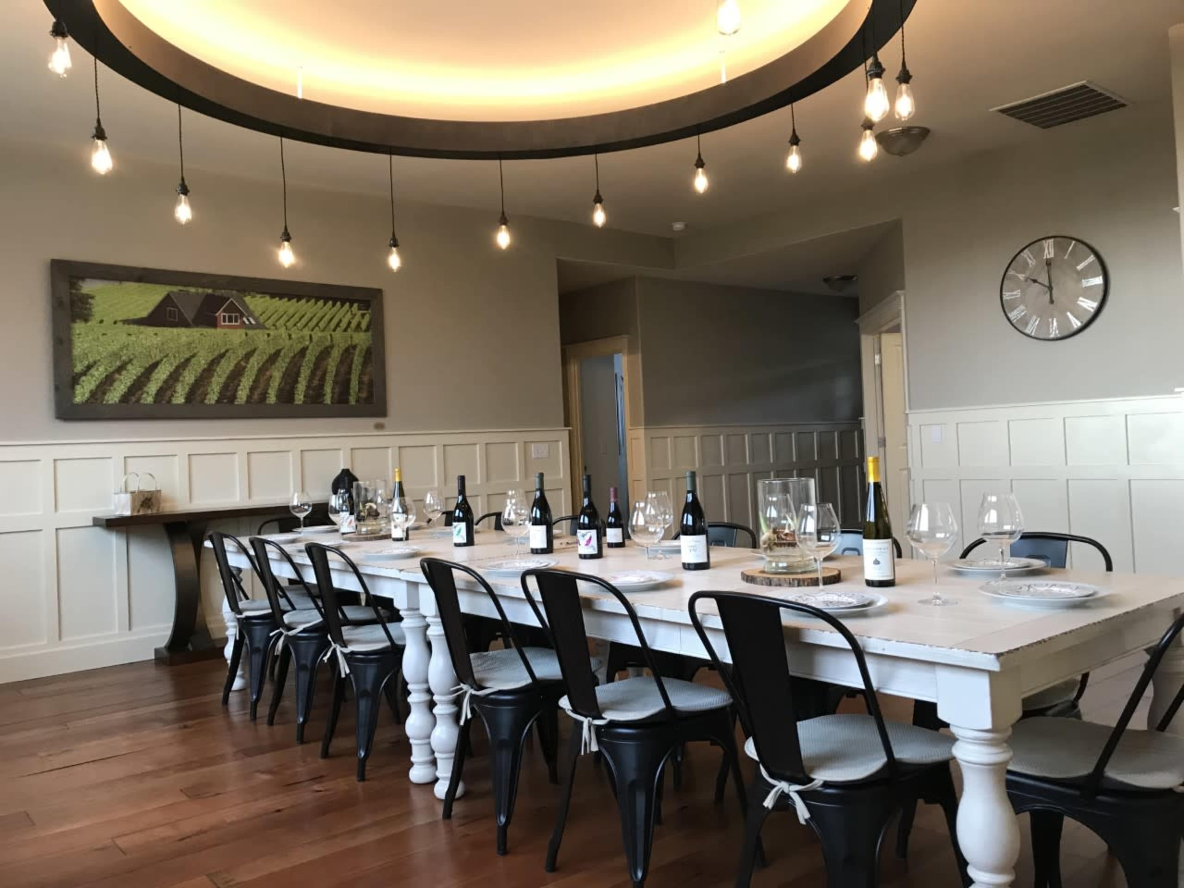 A long dining table is set with wine bottles and glasses under a circular light fixture, in a room featuring vineyard artwork and wooden flooring.