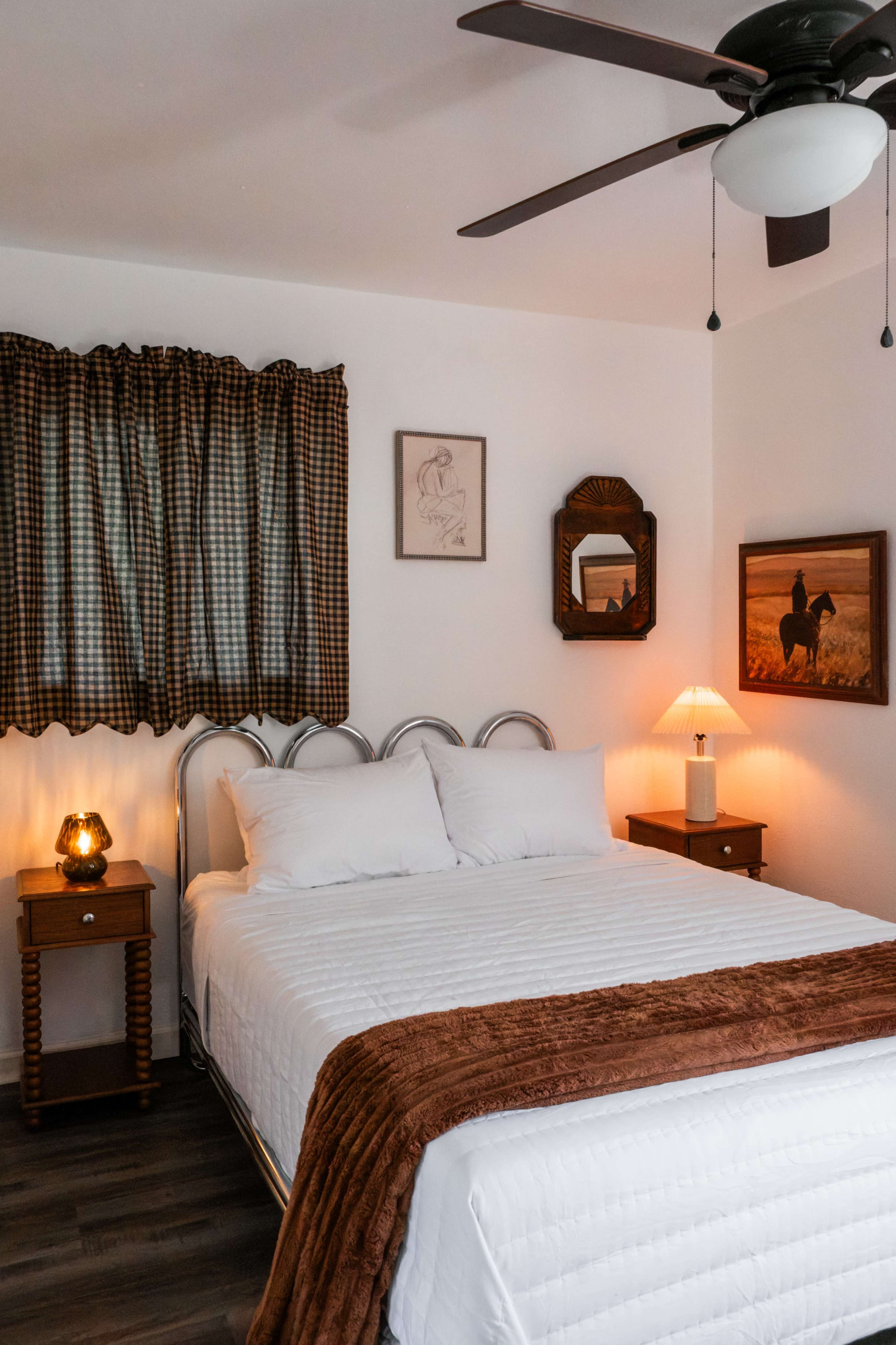 A neatly arranged bedroom features a large bed with white linens, wooden nightstands, a ceiling fan, and warm lighting from a lamp and a decorative lamp on the nightstand.