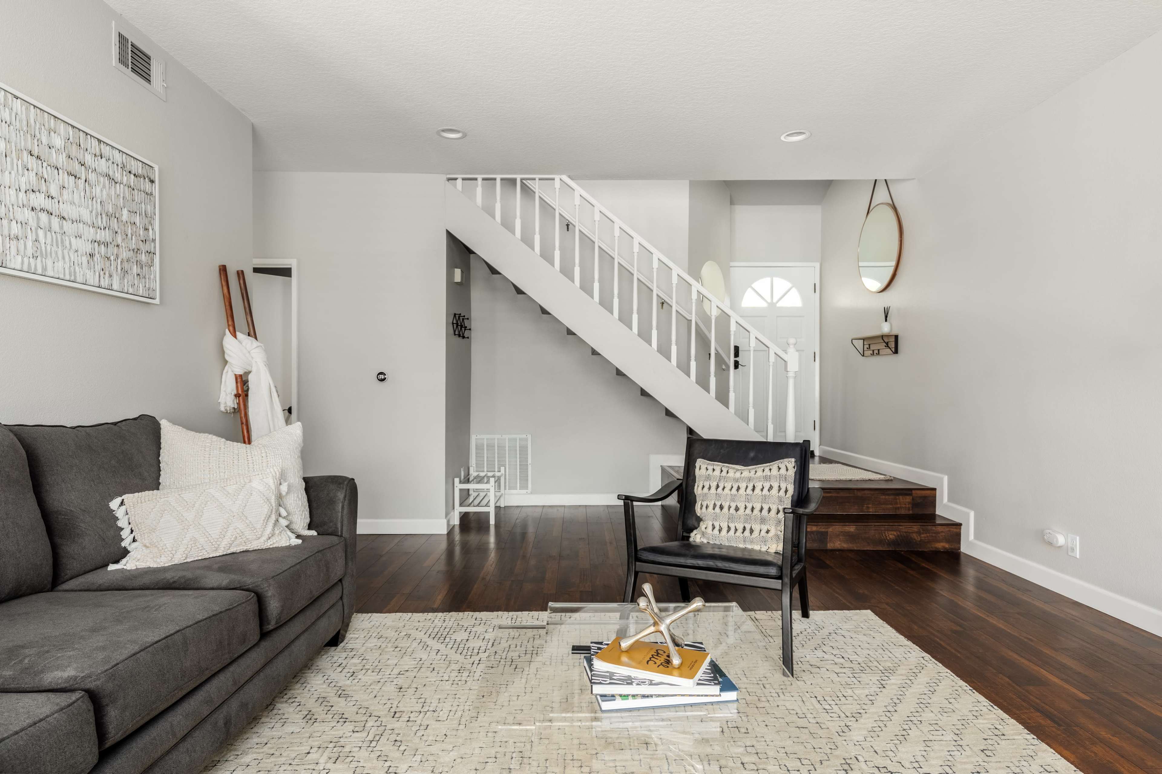 The image shows a modern living room with a gray sofa, a decorative rug, and a staircase leading to the upper level.