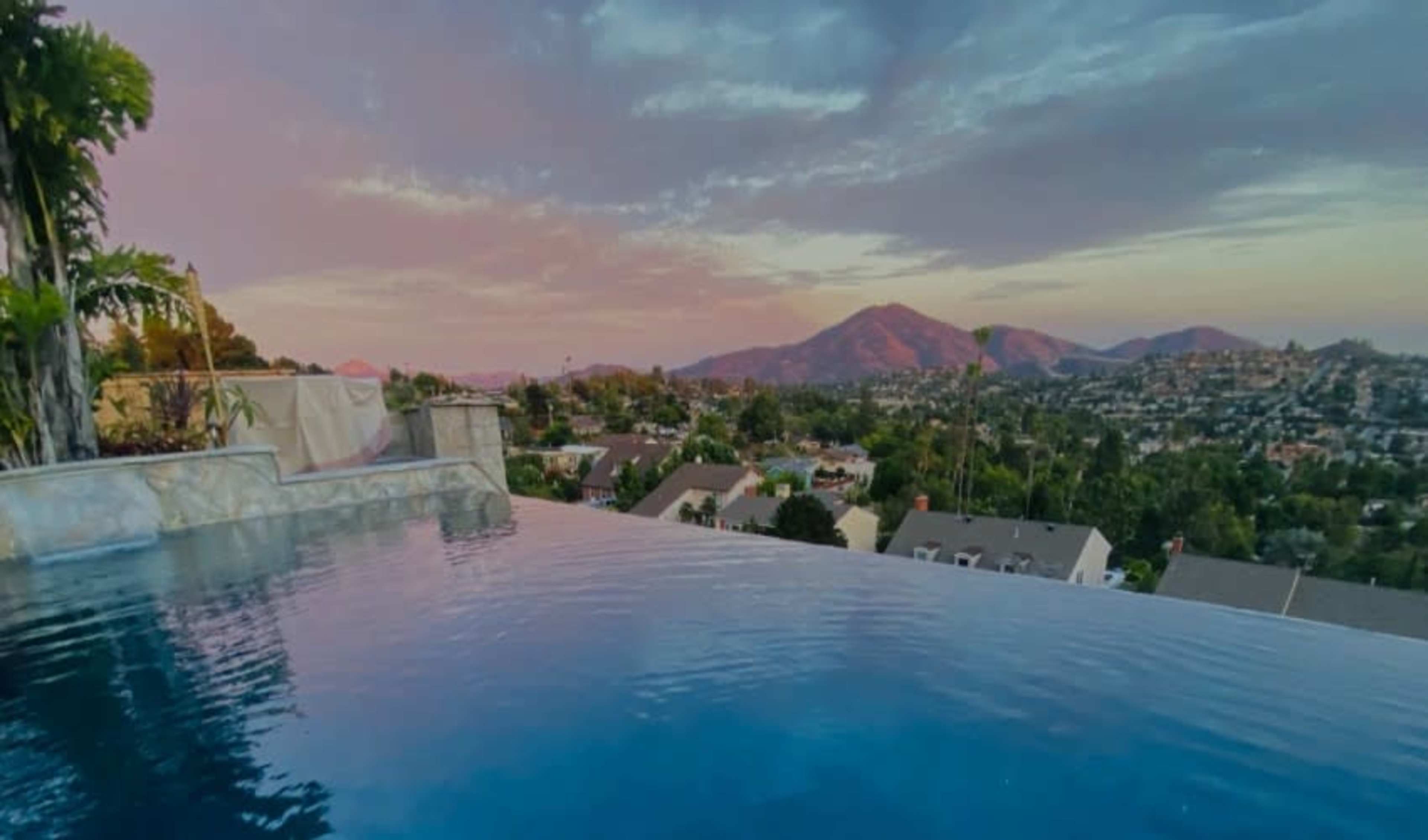 An infinity pool overlooking a residential area and mountains at sunset.