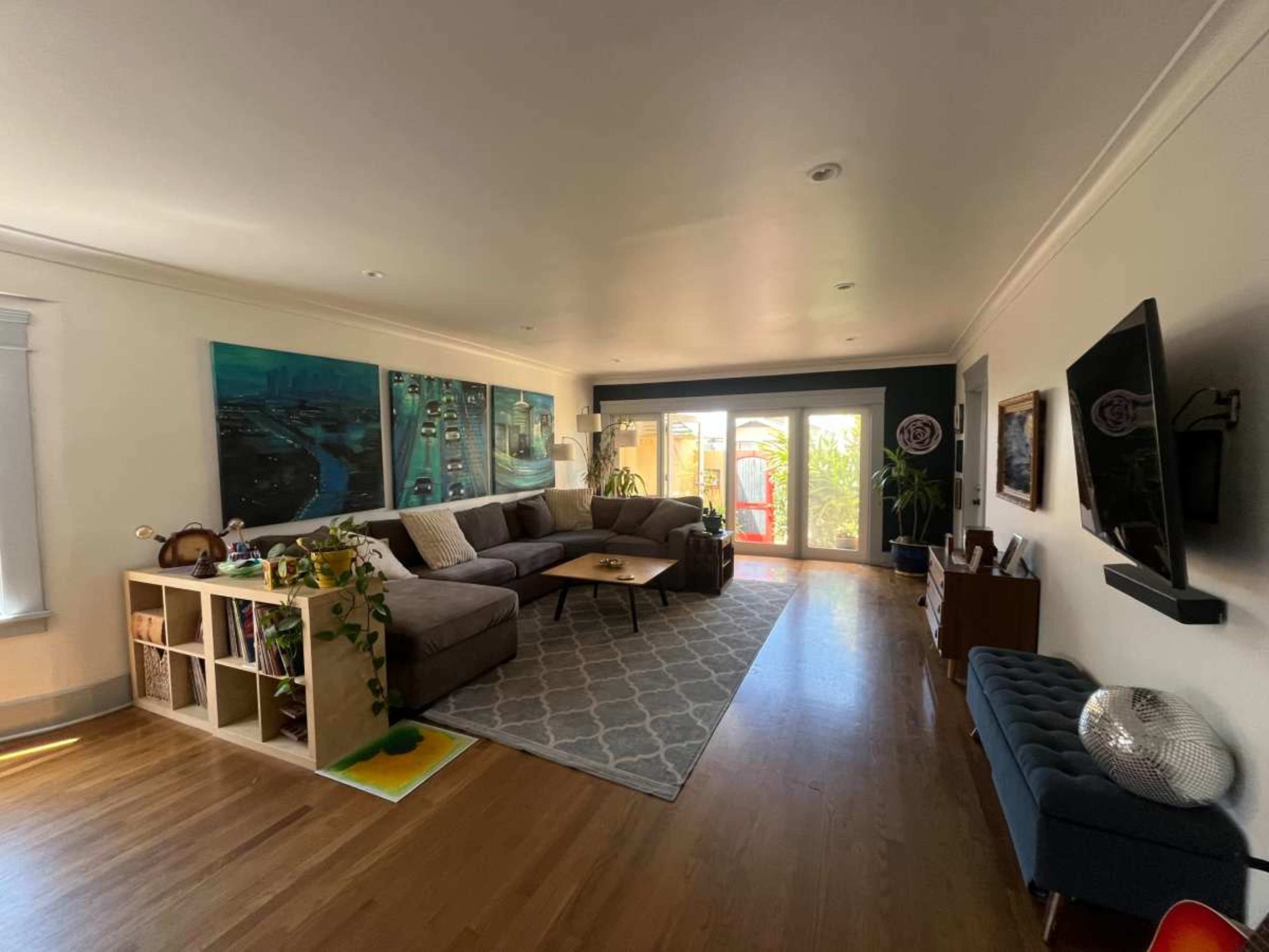 The image shows a spacious living room featuring a large sectional sofa, a coffee table, and bright artwork on the walls, with large windows overlooking a patio.