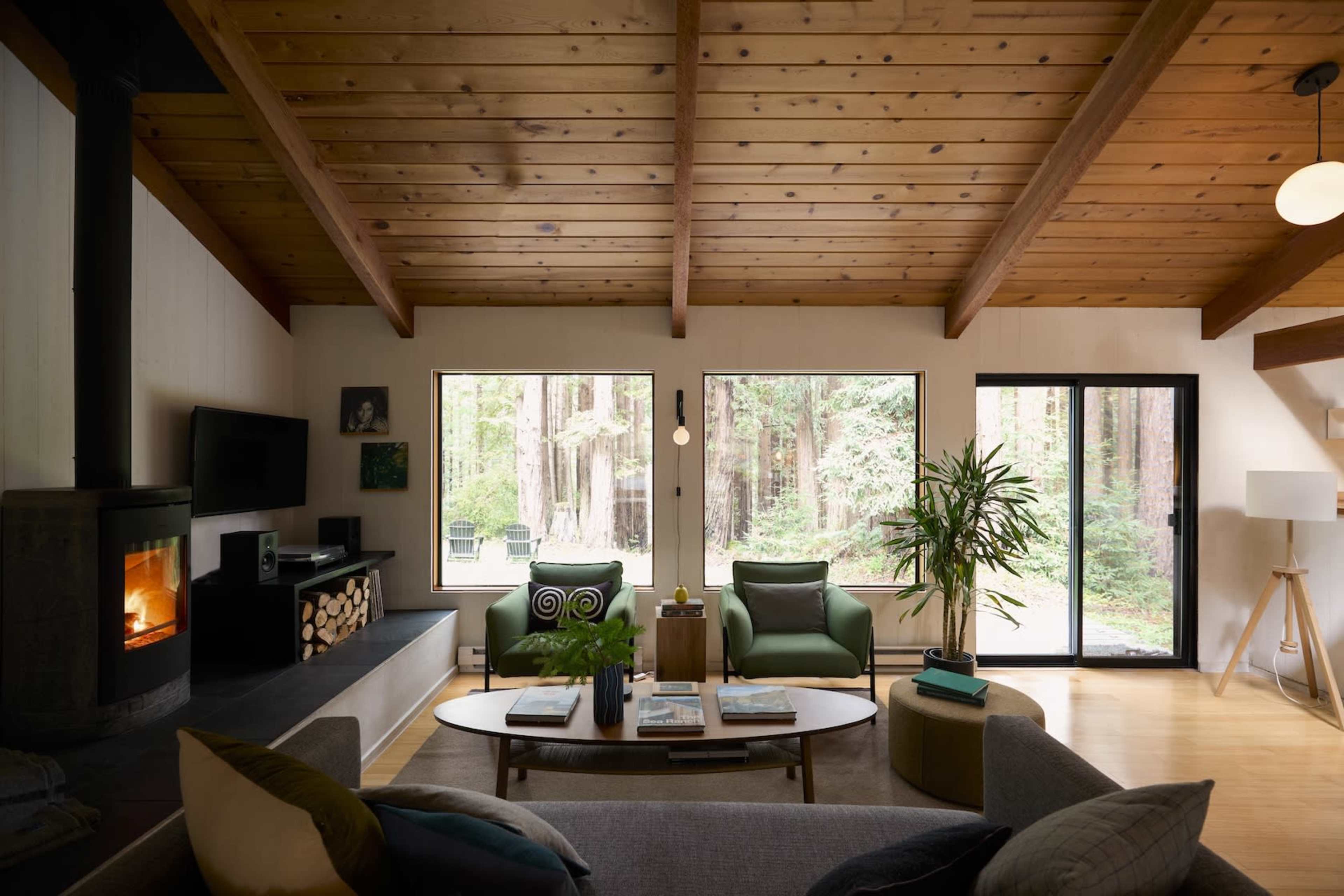 A modern living room features large windows overlooking a forest, with a couch, two chairs, a coffee table, and a wood stove.
