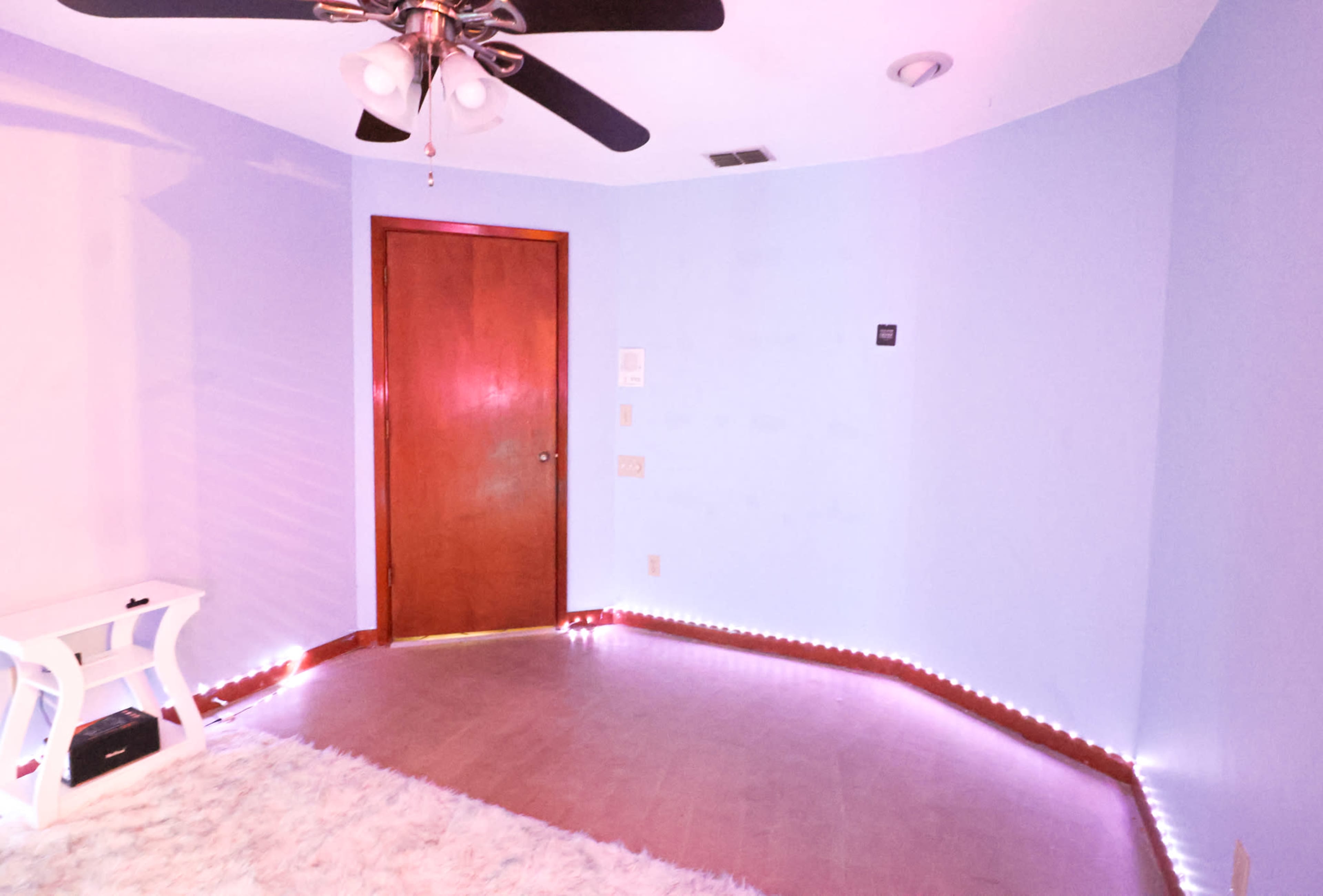 The image shows a sparsely furnished room with pale blue walls, a wooden door, and LED strip lighting along the floor.