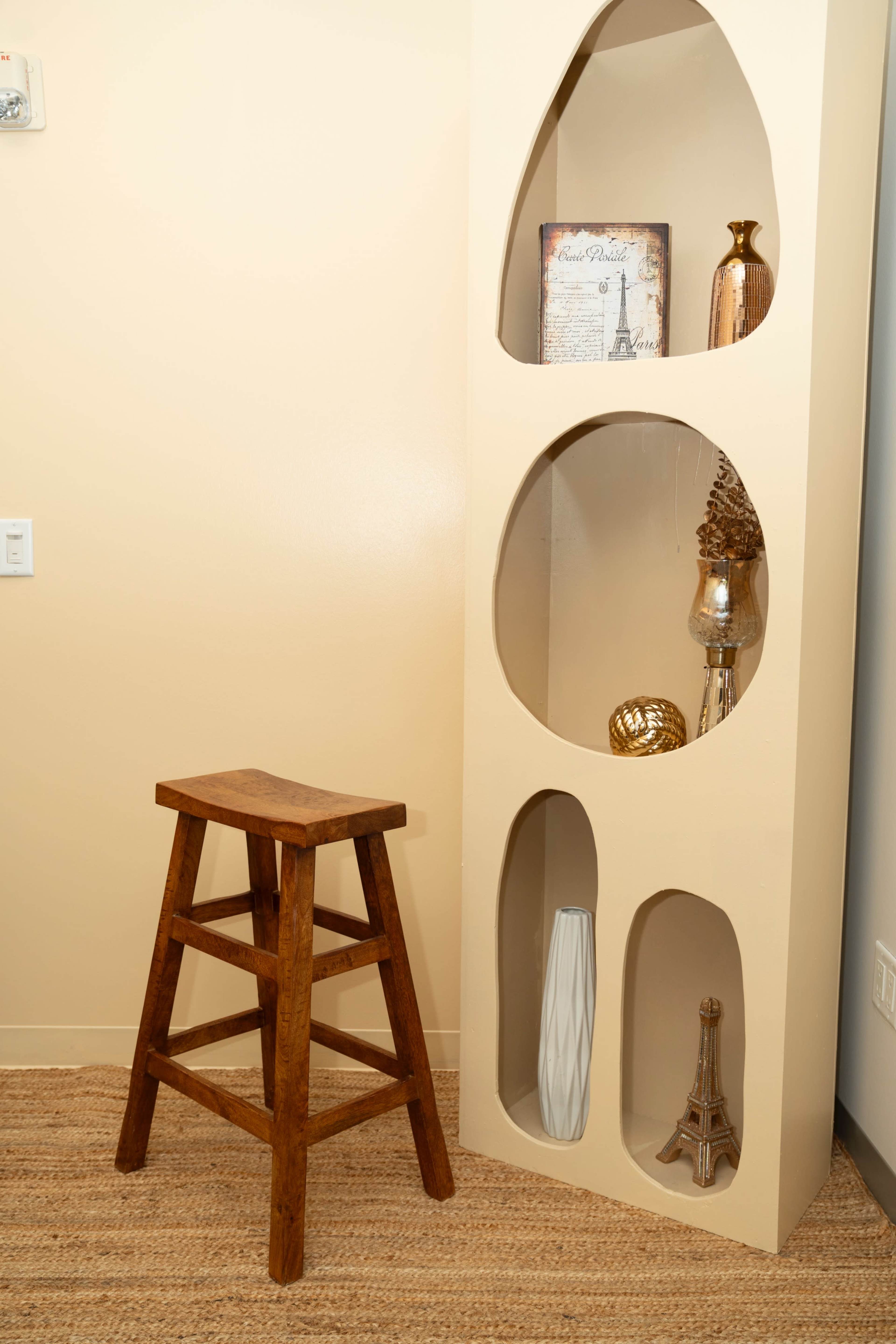 A wooden stool stands next to a beige display unit with circular cutouts that hold decorative items and books.
