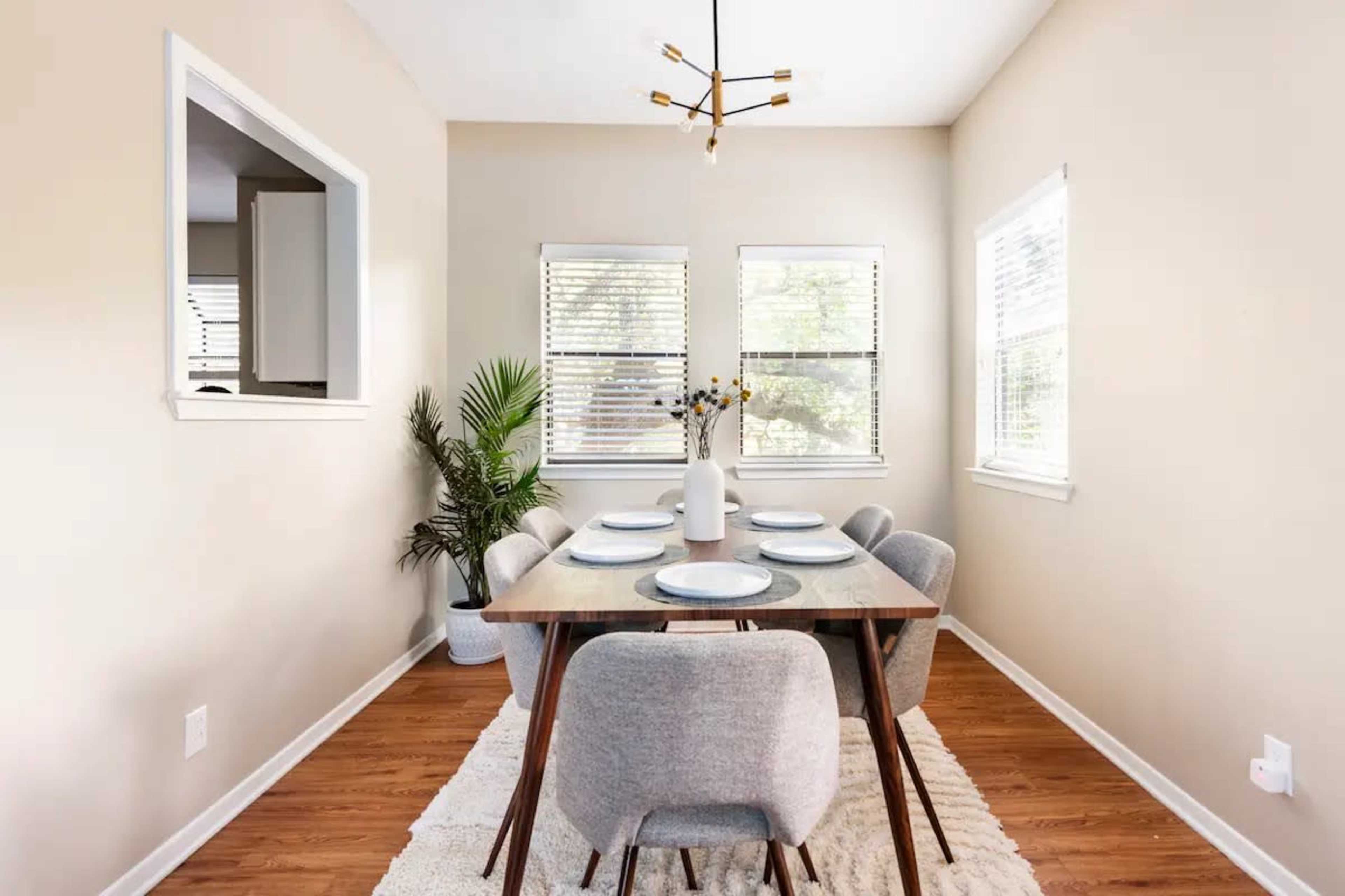 A dining area features a wooden table set for four, surrounded by upholstered chairs, with a potted plant in one corner and natural light coming through the windows.