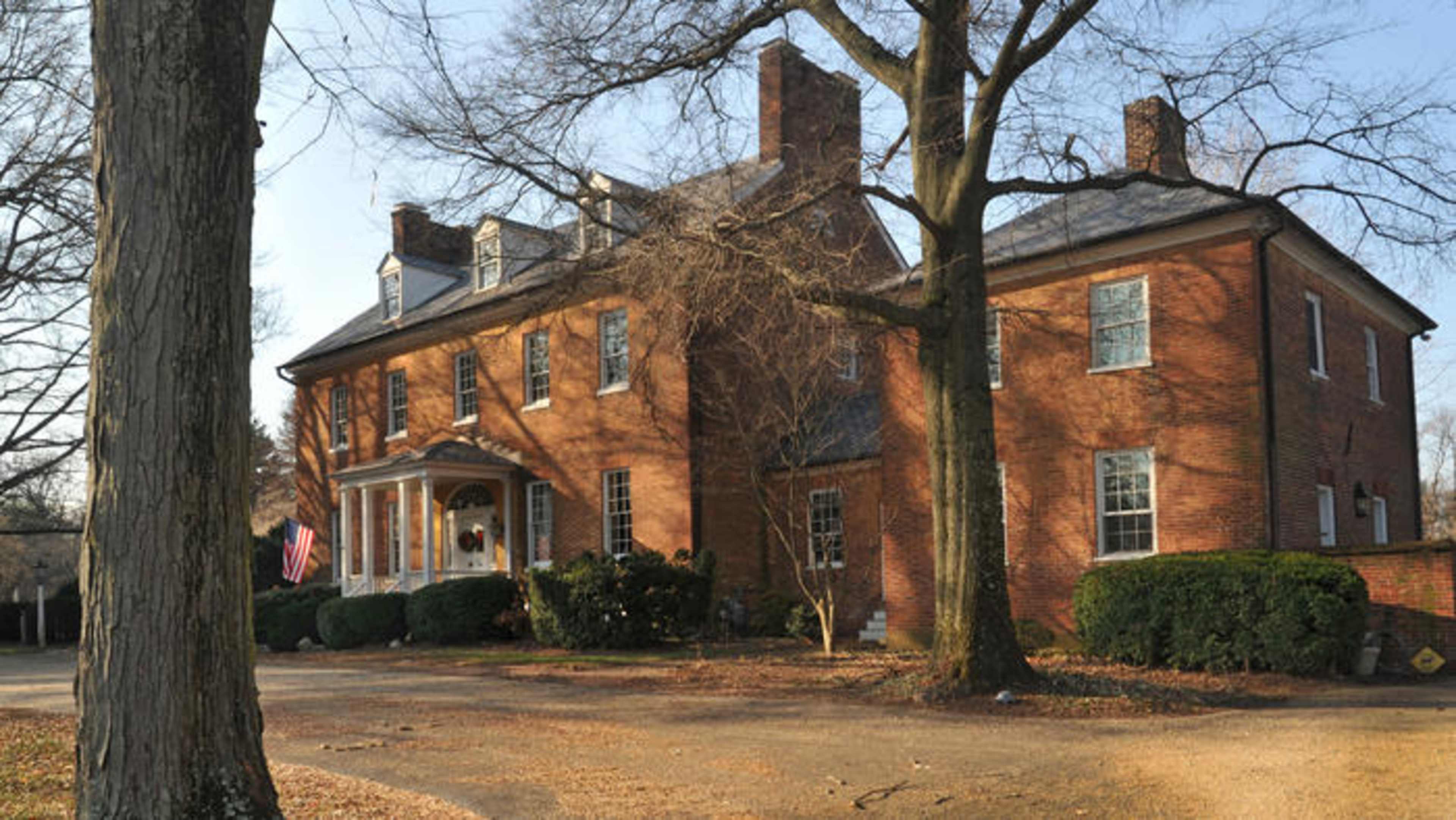 A large, two-story brick house with a covered porch and an American flag, surrounded by trees and trimmed bushes.