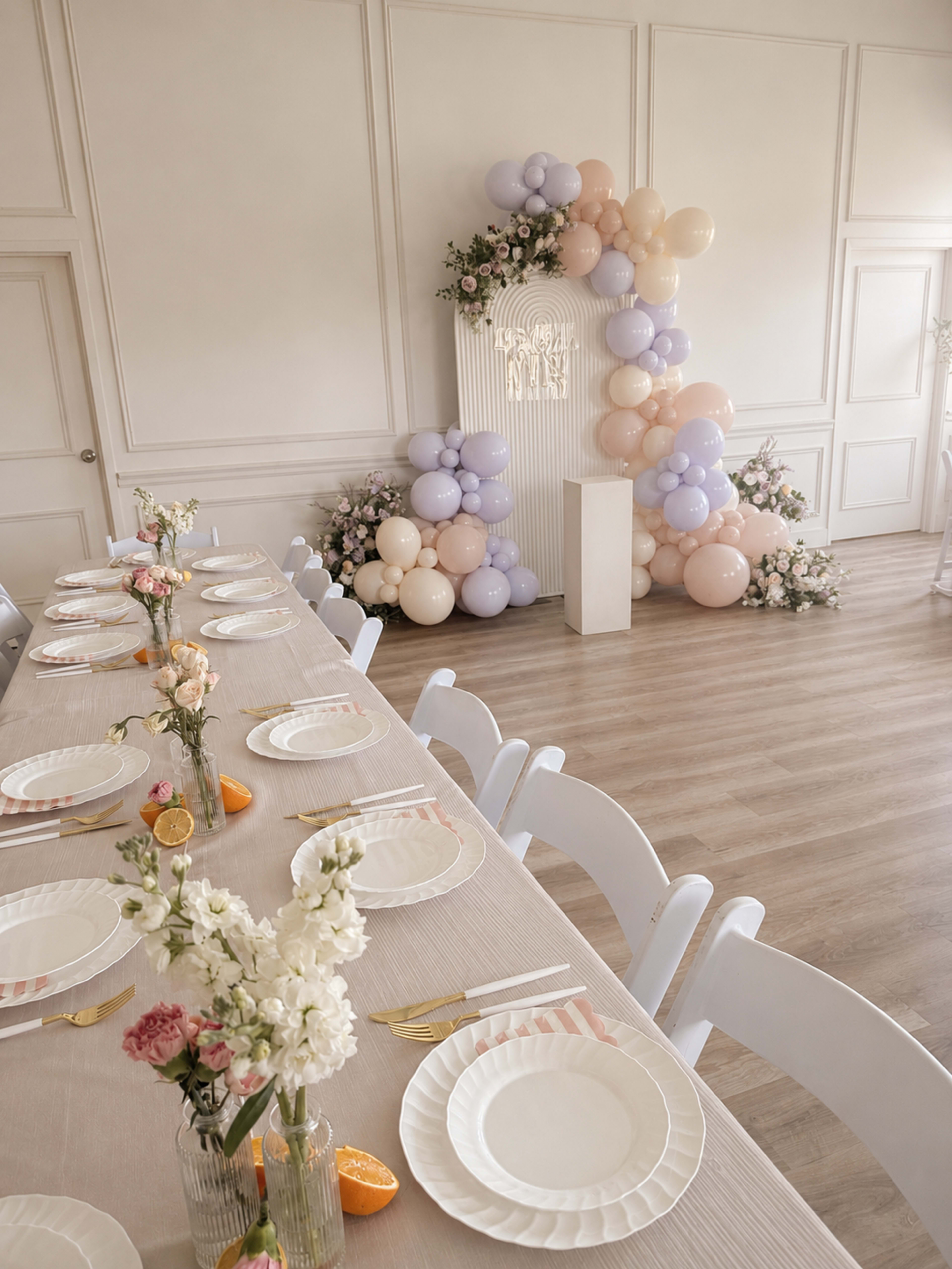 A long dining table is set with white plates and floral arrangements, adjacent to a decorative backdrop of pastel balloons and flowers.