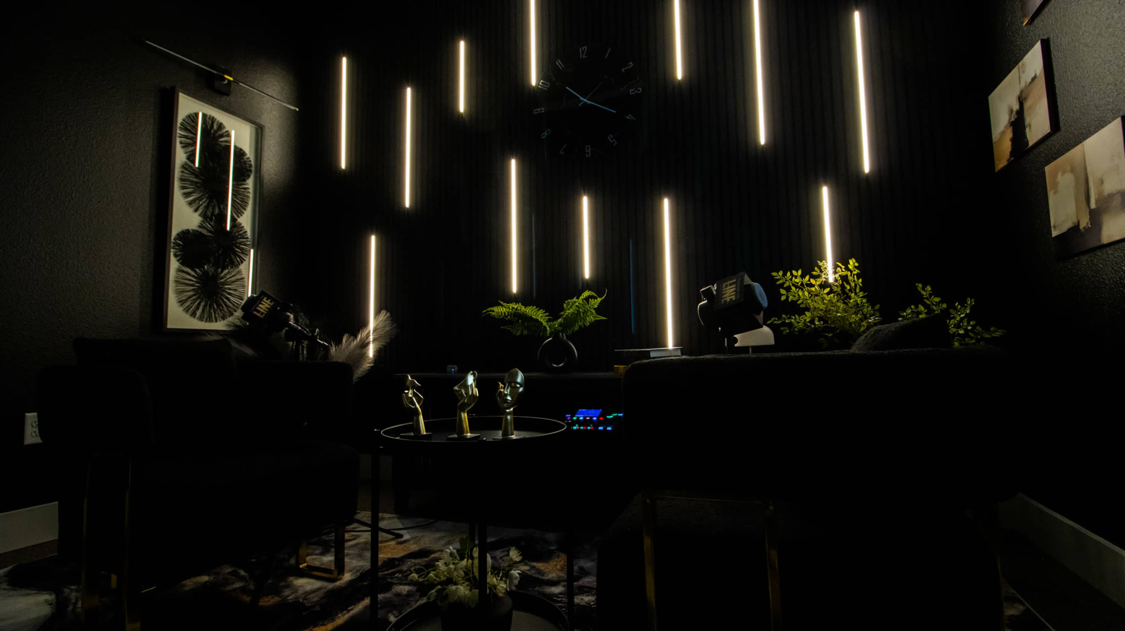 The image features a dimly lit room with vertical light strips, two black chairs, a small round table with glasses, and decorative plants.