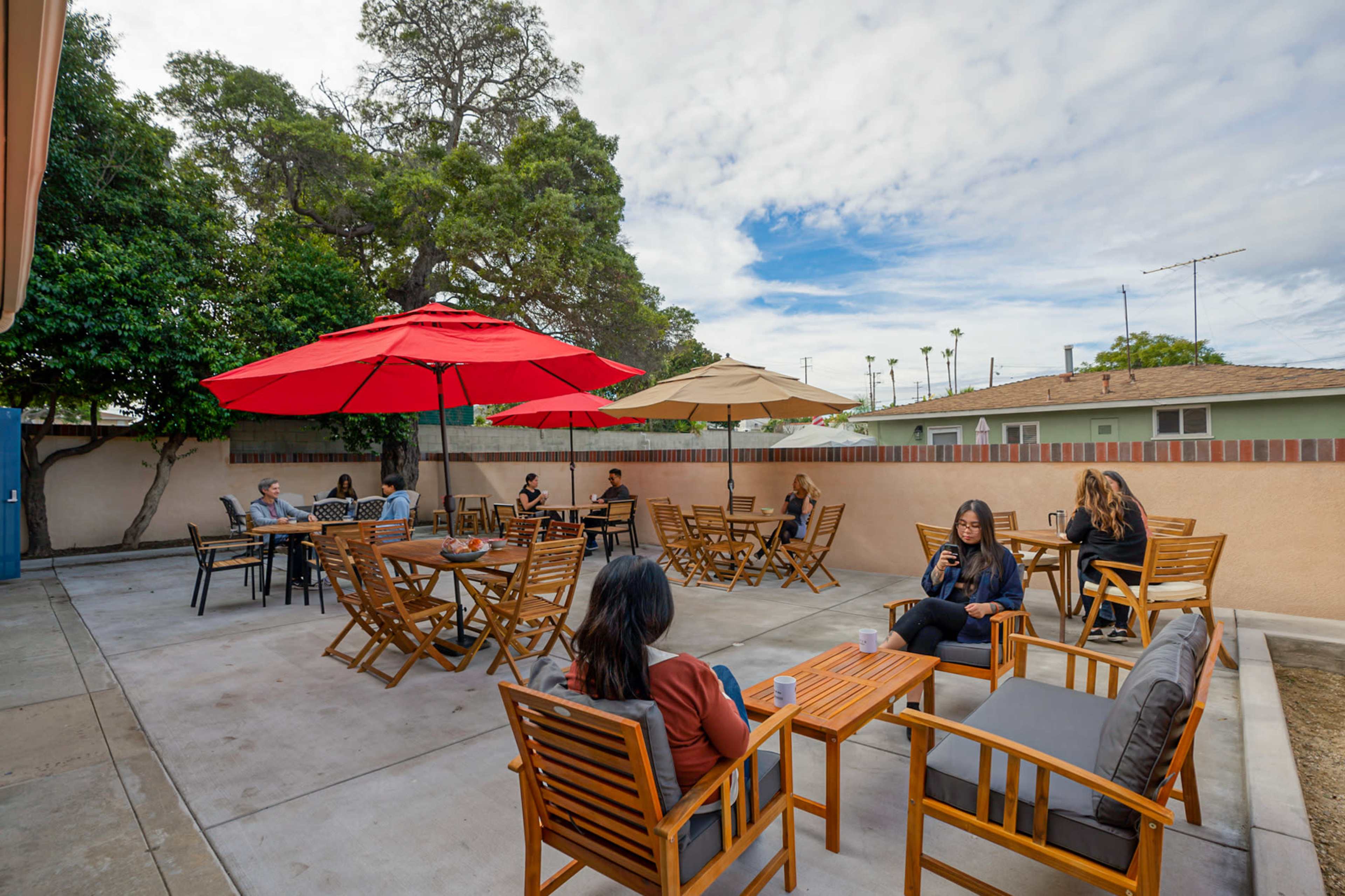 A patio area with several groups of people seated at wooden tables beneath red and beige umbrellas.