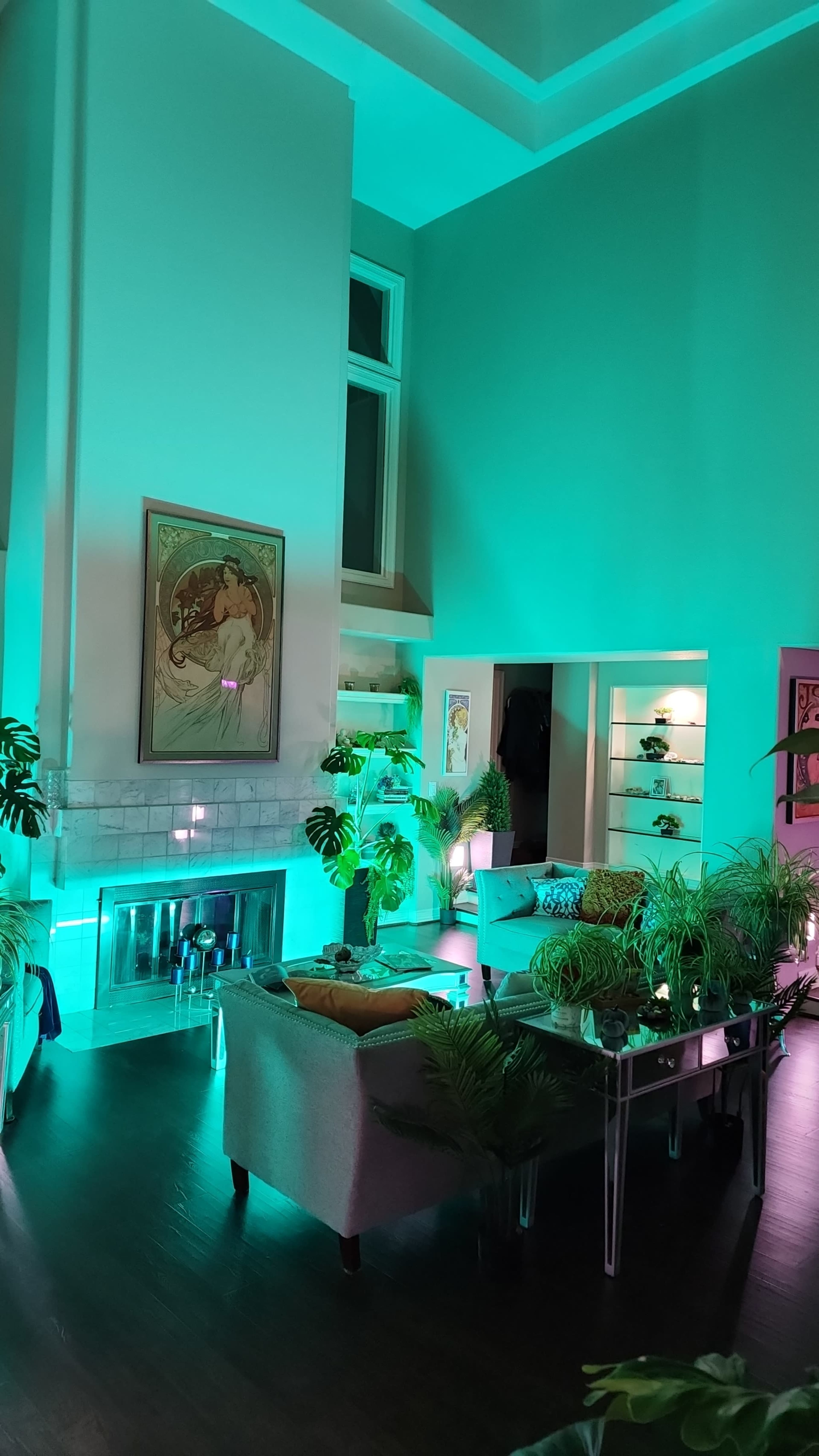 The image shows a modern living room with tall ceilings, featuring a couch, decorative plants, and ambient teal lighting.
