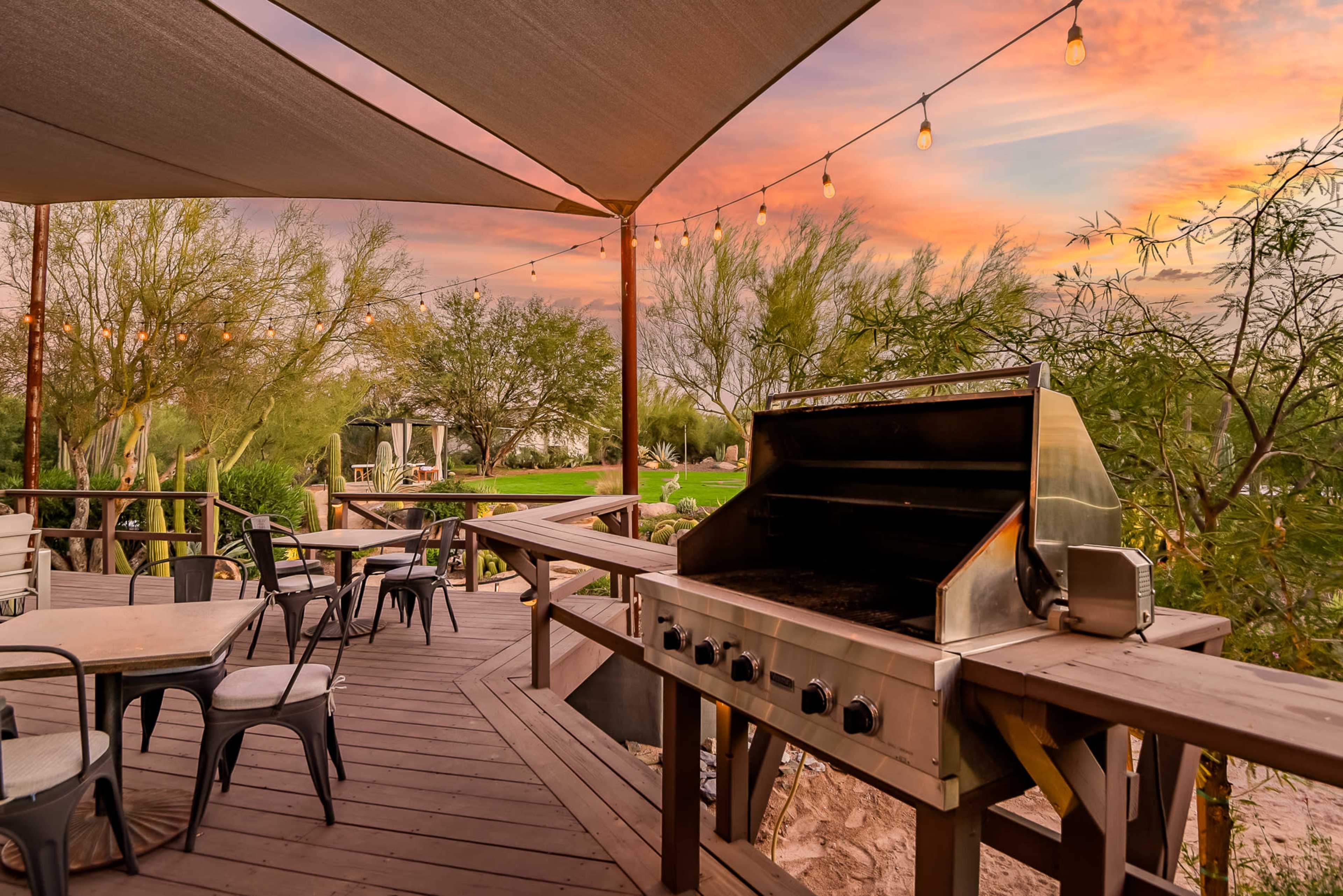 The image shows a wooden deck with an outdoor grill, tables, and string lights, set against a colorful sunset sky.